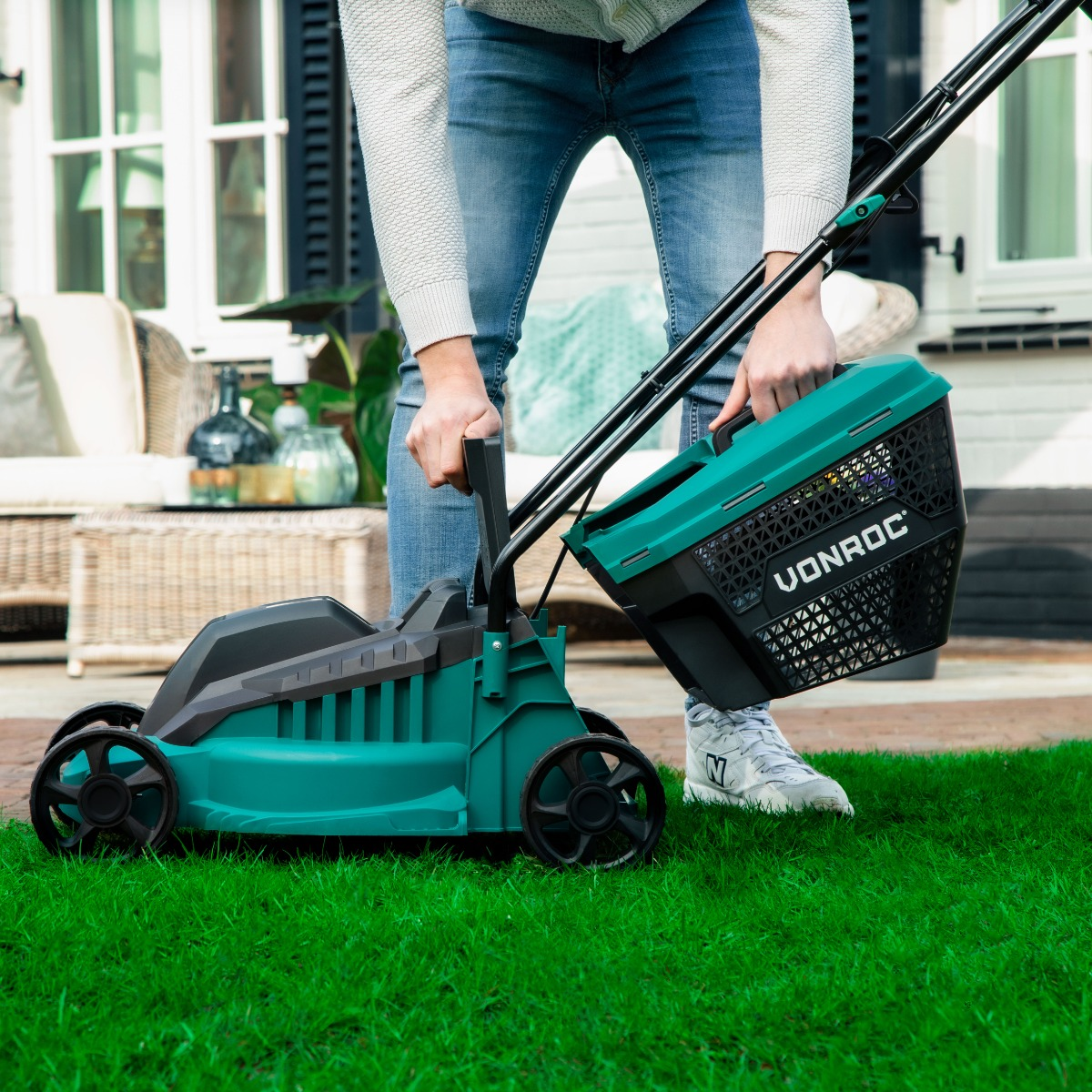 Persoon houdt een groene grasmaaier met een zwarte mand vast. Het gazon is groen. De achtergrond is een huis.