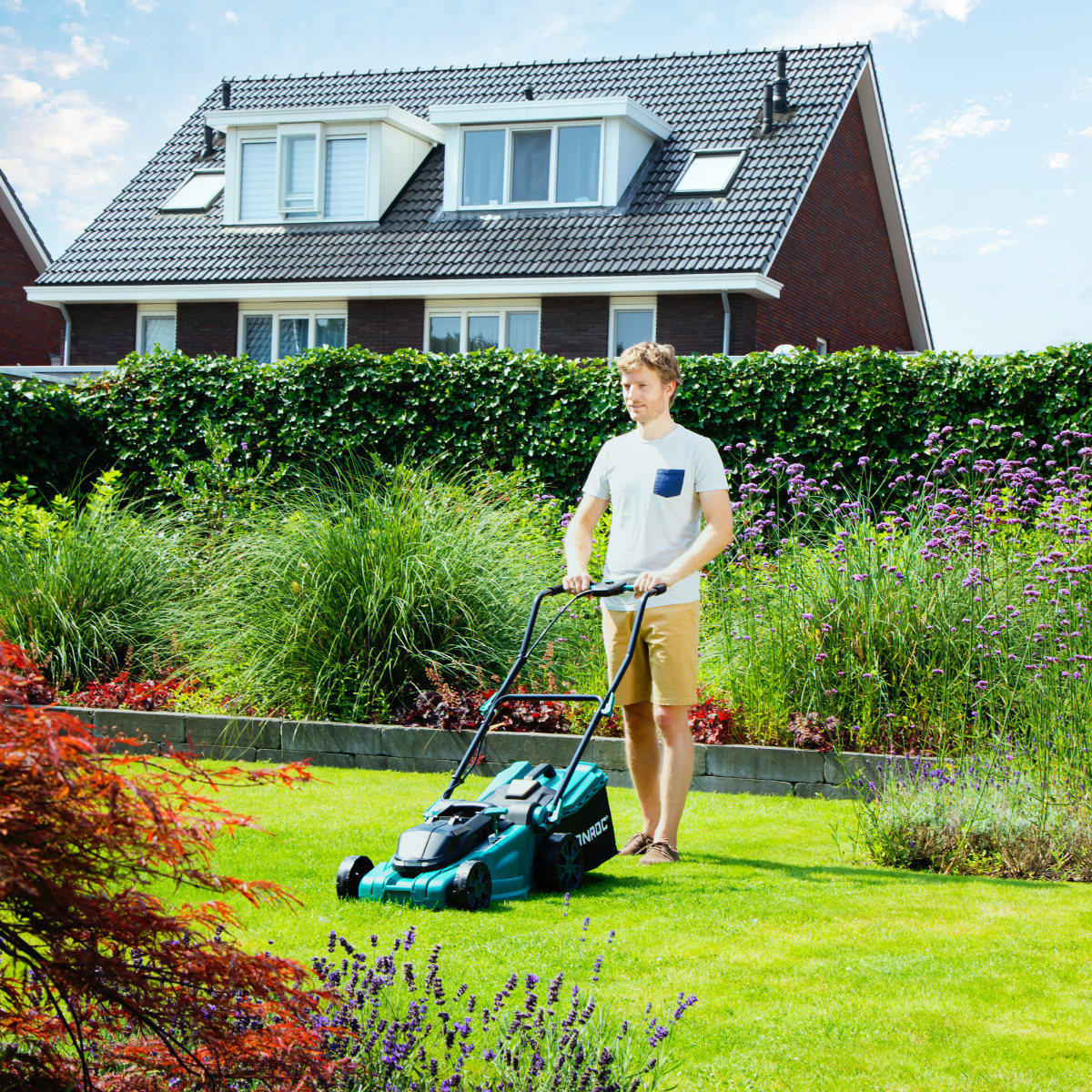Een man maait een groen gazon met een turquoise grasmaaier. Op de achtergrond staat een huis met een donker dak. Struiken en bloemen omringen het gazon.