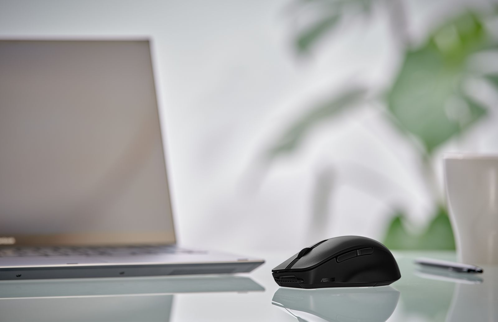 Un mouse nero per computer si trova su una superficie di vetro. Sono visibili anche un laptop, una tazza bianca e una penna.