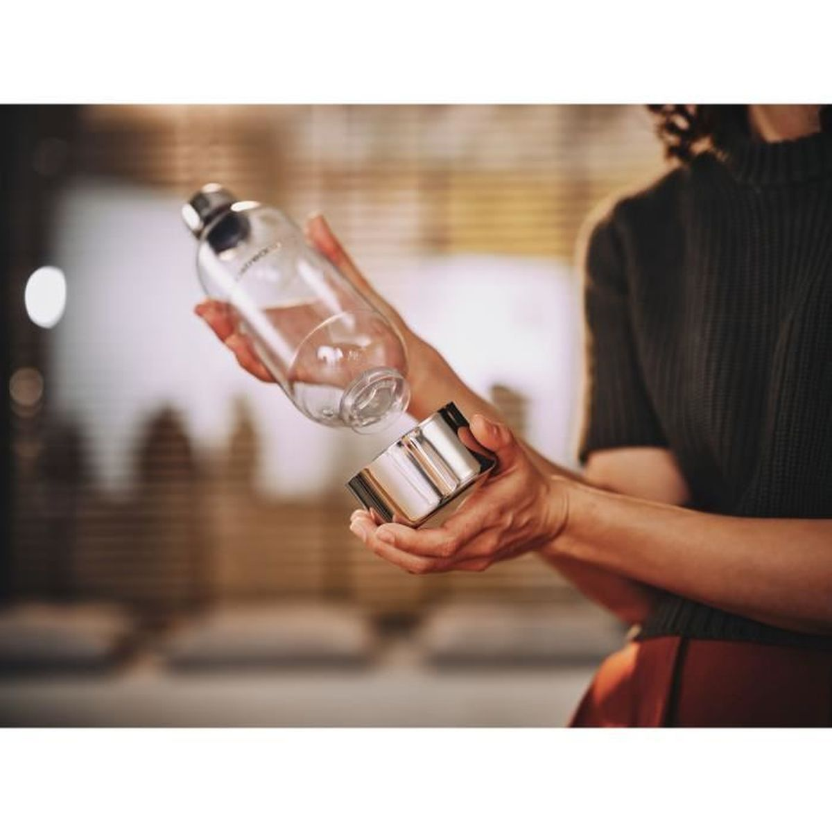 Eine Person hält eine klare Flasche über einem silbernen Becher und bereitet das Eingießen vor. Der Hintergrund ist unscharf, mit etwas Licht.