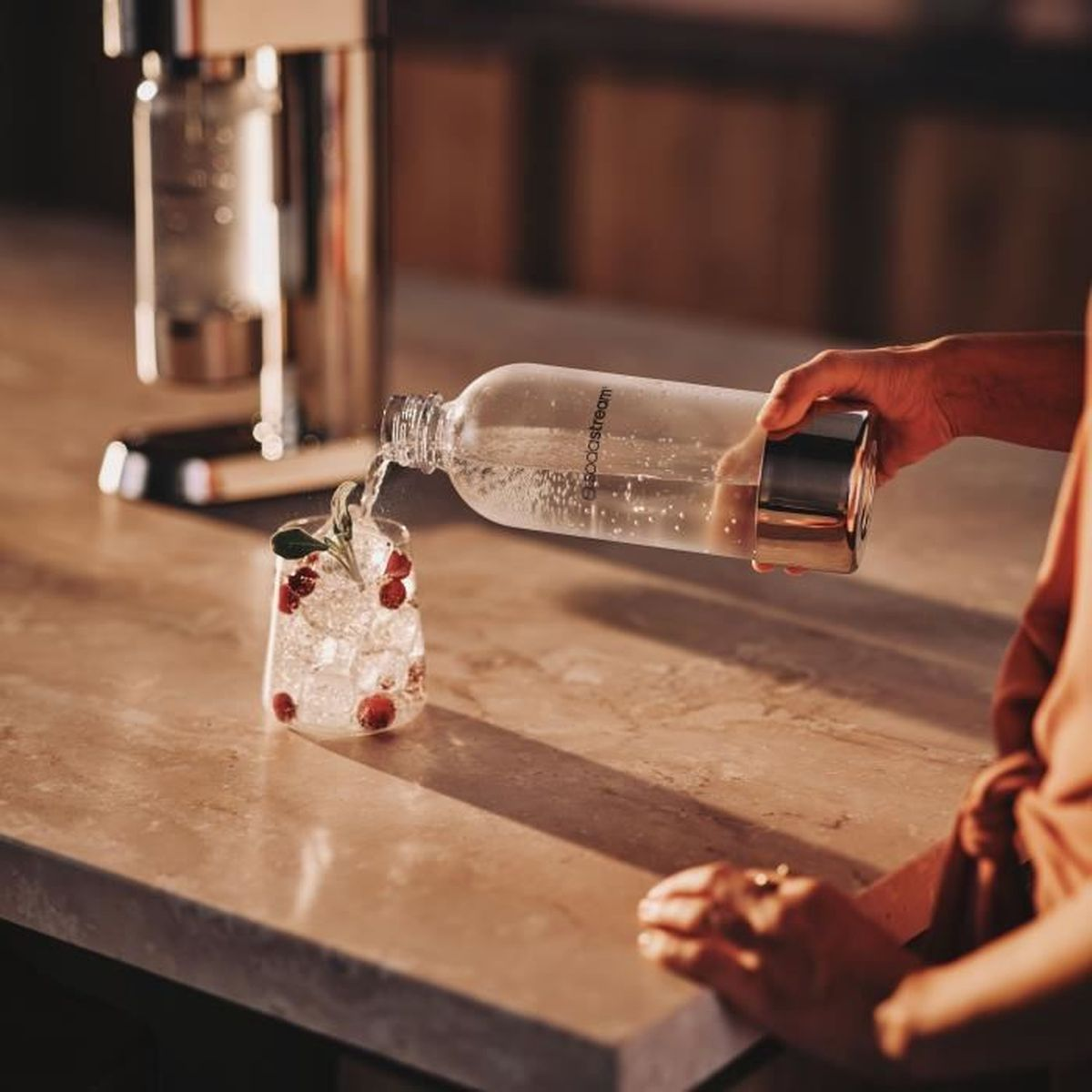 Eine Person gießt Wasser aus einer Flasche in ein Glas mit Eis, Preiselbeeren und einem Zweig Grünzeug. Im Hintergrund ist ein Sodabereiter zu sehen.