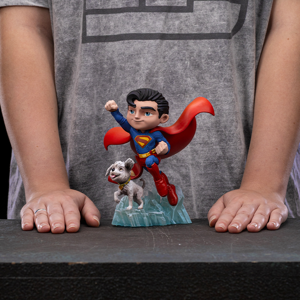 Una persona tiene una statuetta di Superman con un cane. Superman vola con un mantello rosso. Il cane è bianco. Lo sfondo è grigio.