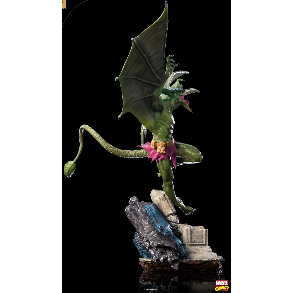 Una statua verde a forma di drago con grandi ali e una lunga coda, in posa su una base di pietra, su sfondo nero.