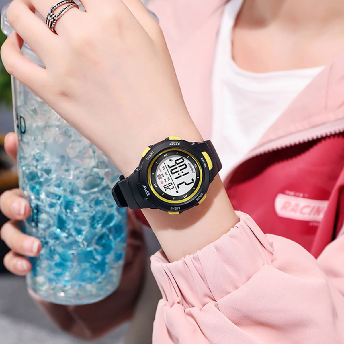 Eine Person trägt eine schwarz-gelbe Digitaluhr. Sie hält ein Getränk und trägt eine rosa Jacke. Ein Ring ist an ihrem Finger.