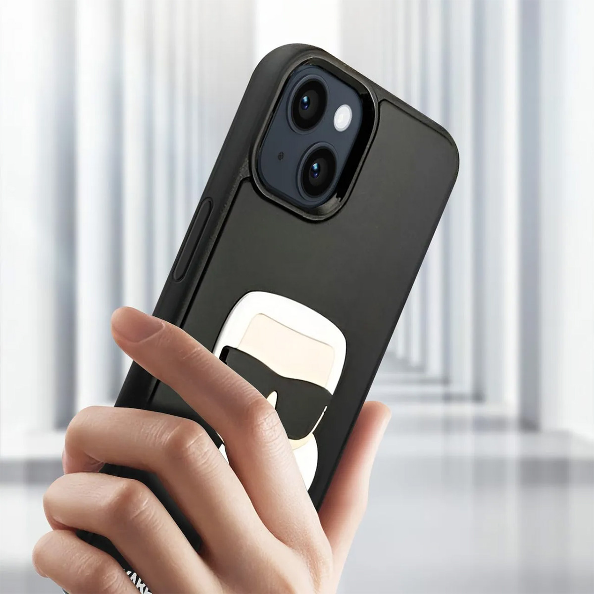 Una mano che tiene un telefono nero con una custodia Karl Lagerfeld. Lo sfondo è uno spazio sfocato, bianco e moderno.