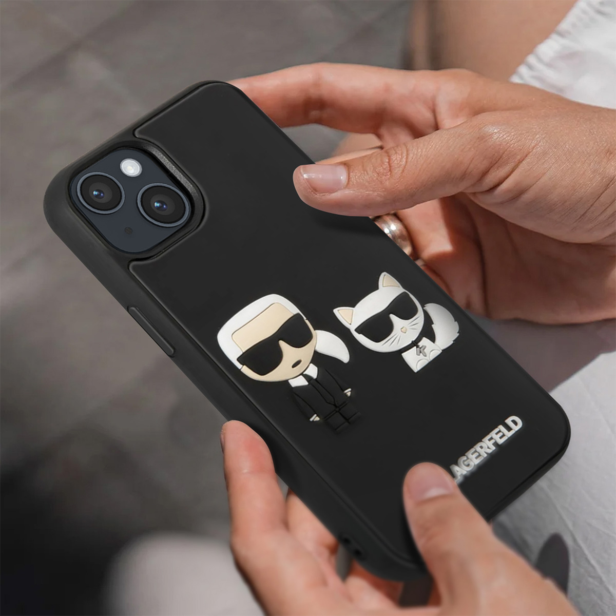 Una persona tiene in mano una custodia nera per telefono con un design a cartoni animati. Il design include un personaggio e un gatto, entrambi con occhiali da sole.