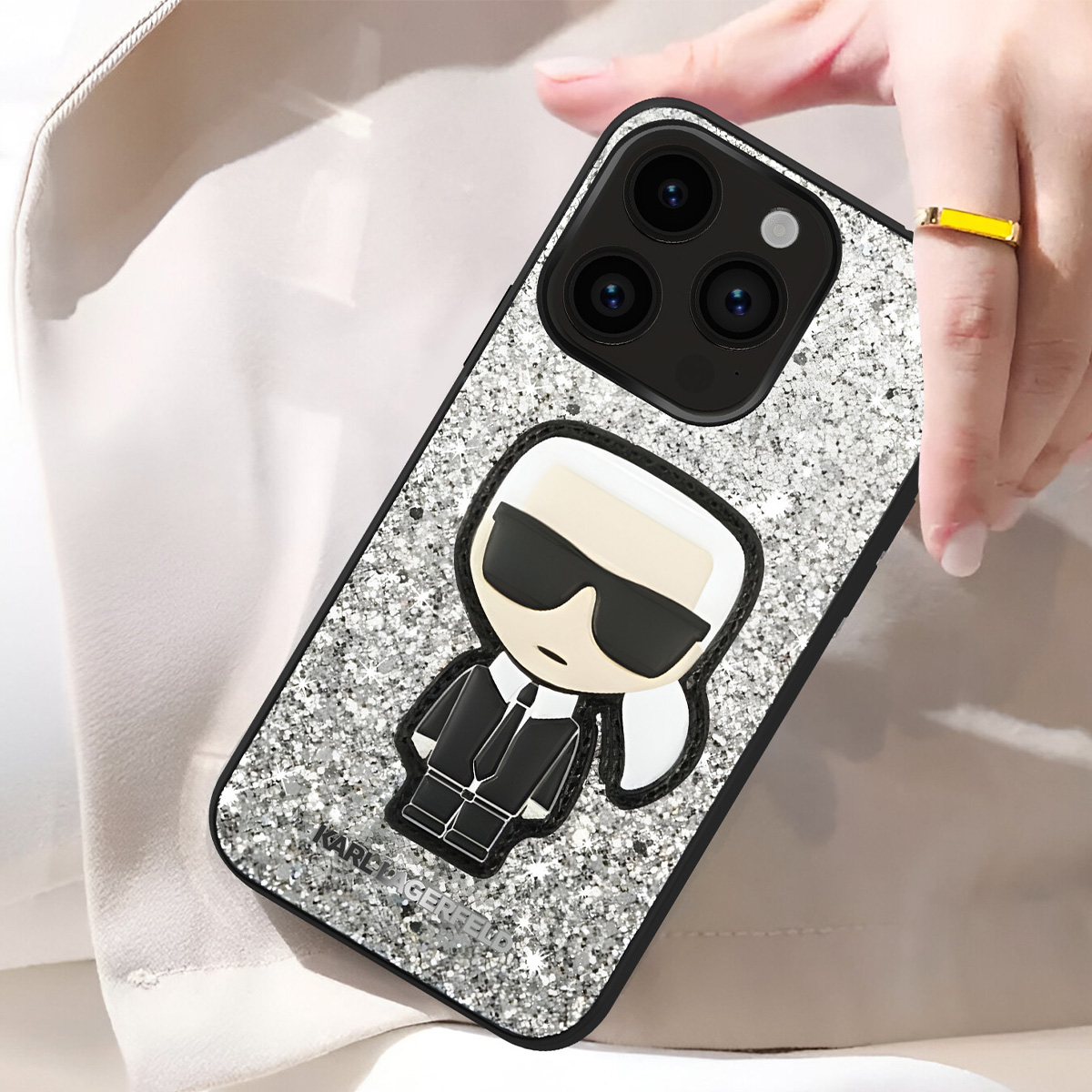 Una custodia per telefono con un design Karl Lagerfeld è tenuta in mano. La custodia è argento glitterato con contorno nero e un personaggio dei cartoni animati.