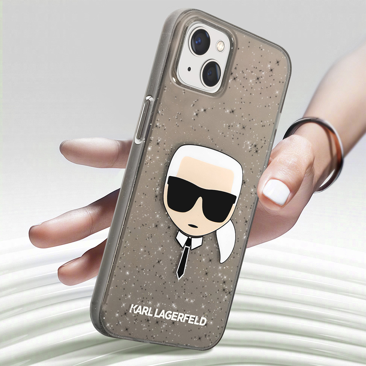 Una mano tiene un telefono con una custodia Karl Lagerfeld. La custodia è grigia con stelle e un cartone di Karl. Sfondo sfocato.