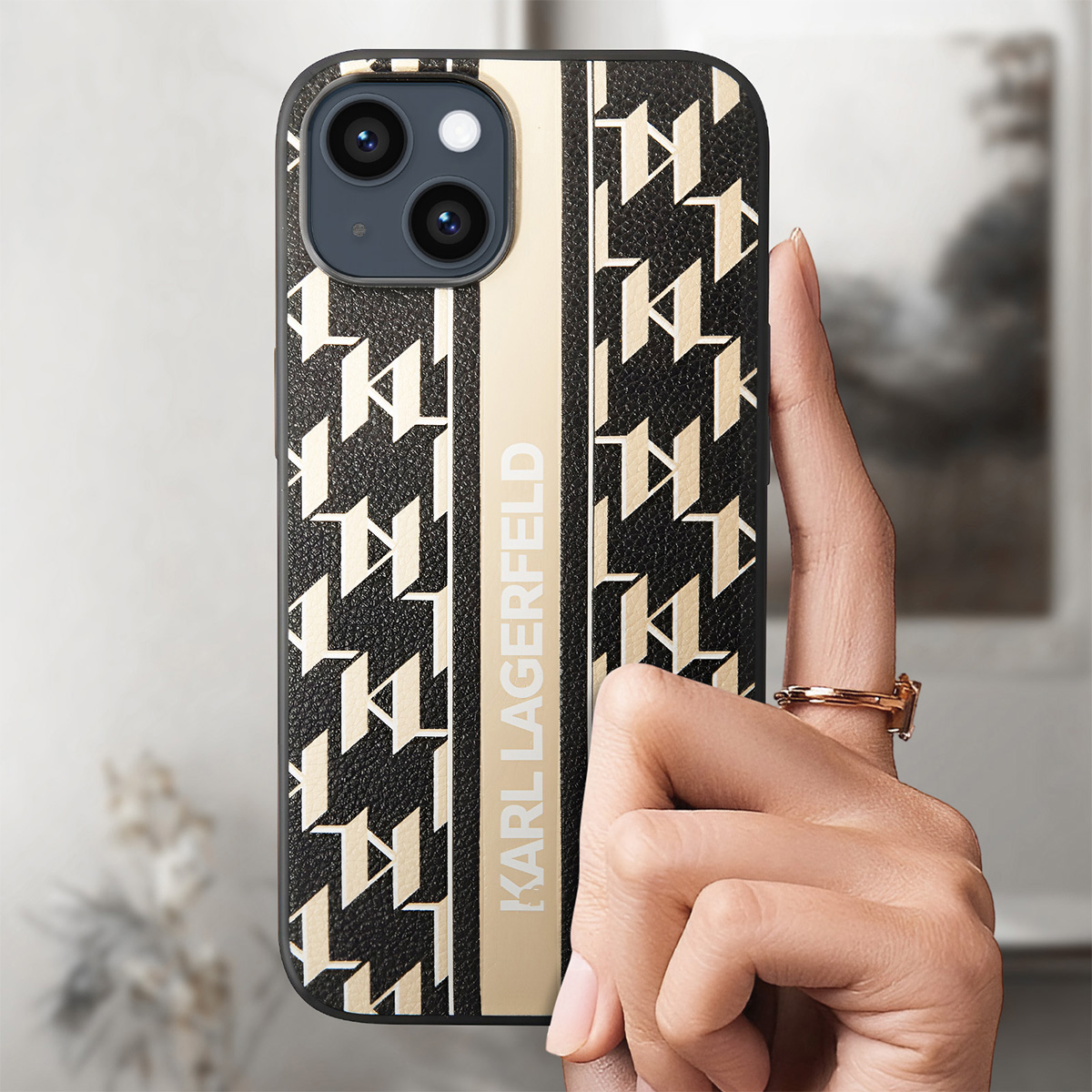 Una mano tiene un telefono con una custodia Karl Lagerfeld. La custodia ha un motivo nero e beige e il nome del marchio. Un anello al dito.