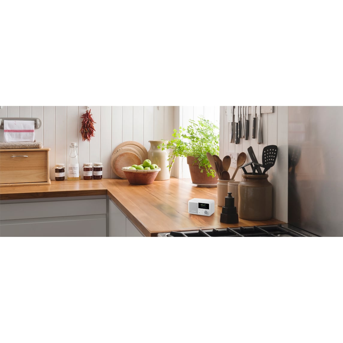A kitchen scene with wooden countertop. A radio sits on the counter. Jars, a bowl of apples, and utensils are also visible.