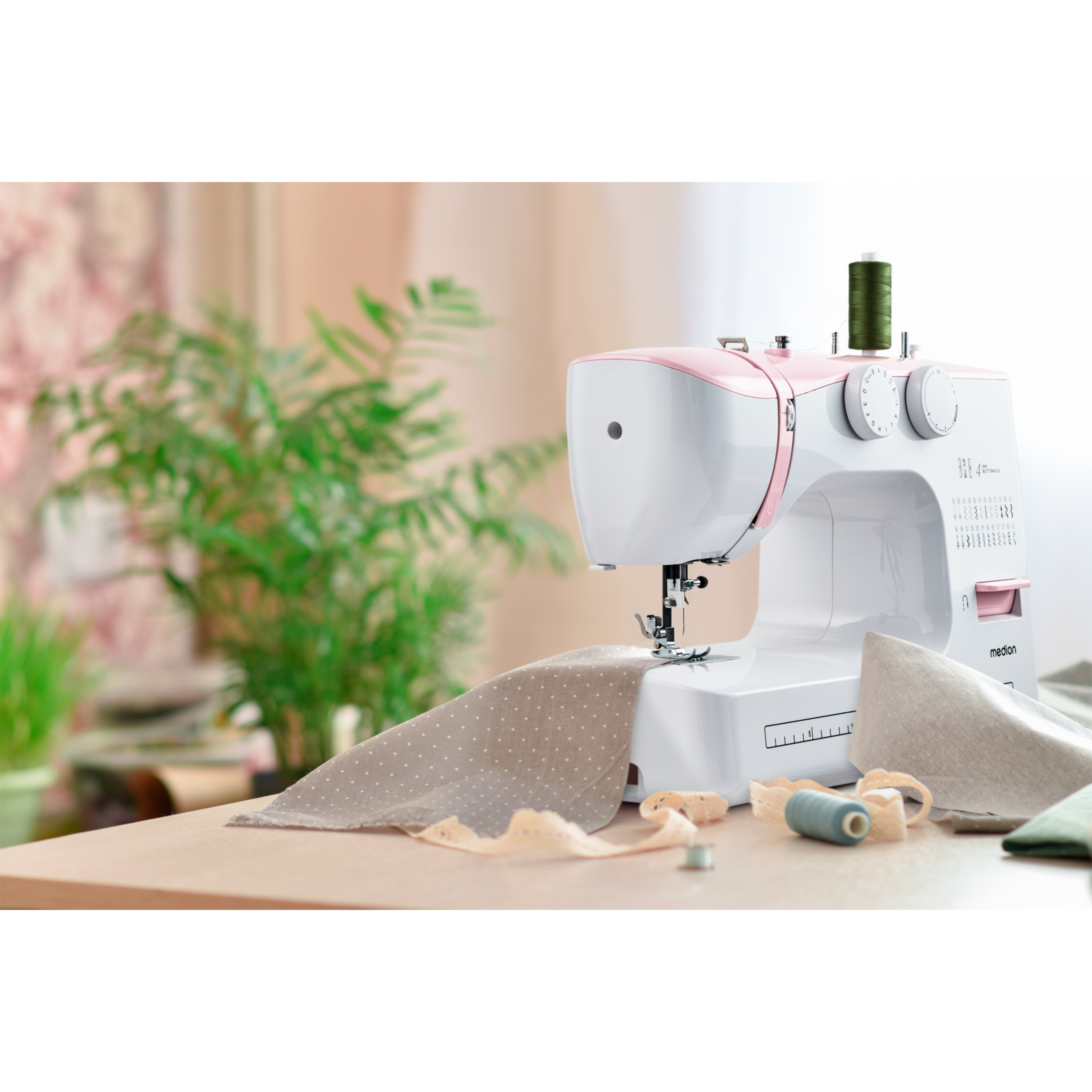 Maszyna do szycia z różowymi akcentami, tkanina, nić i roślina na stole. Tło jest rozmyte.