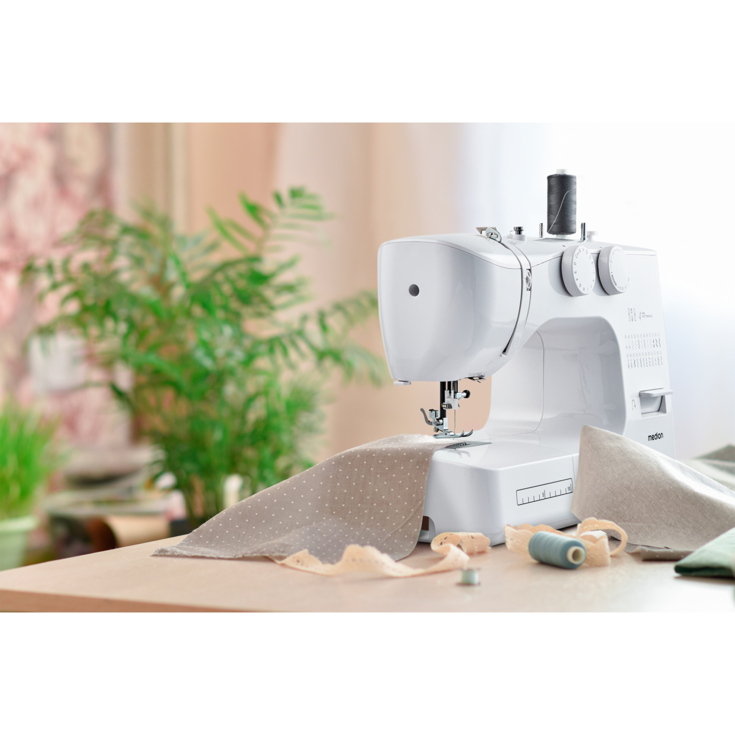 Biała maszyna do szycia z tkaniną i nicią stoi na drewnianym stole, w tle rośliny.