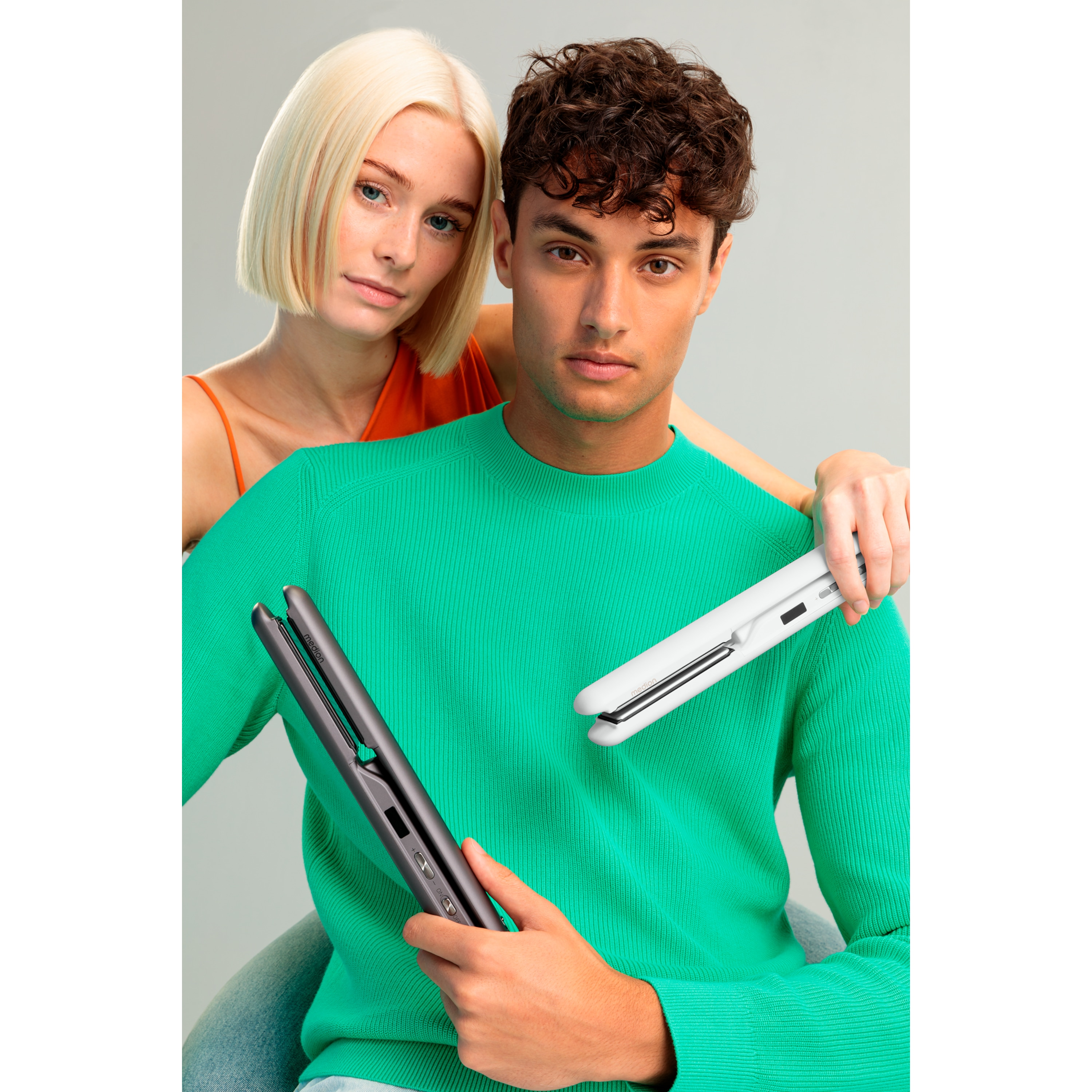 Ein Mann und eine Frau halten Haarglätter. Der Mann trägt Grün, die Frau Orange. Sie schauen in die Kamera.