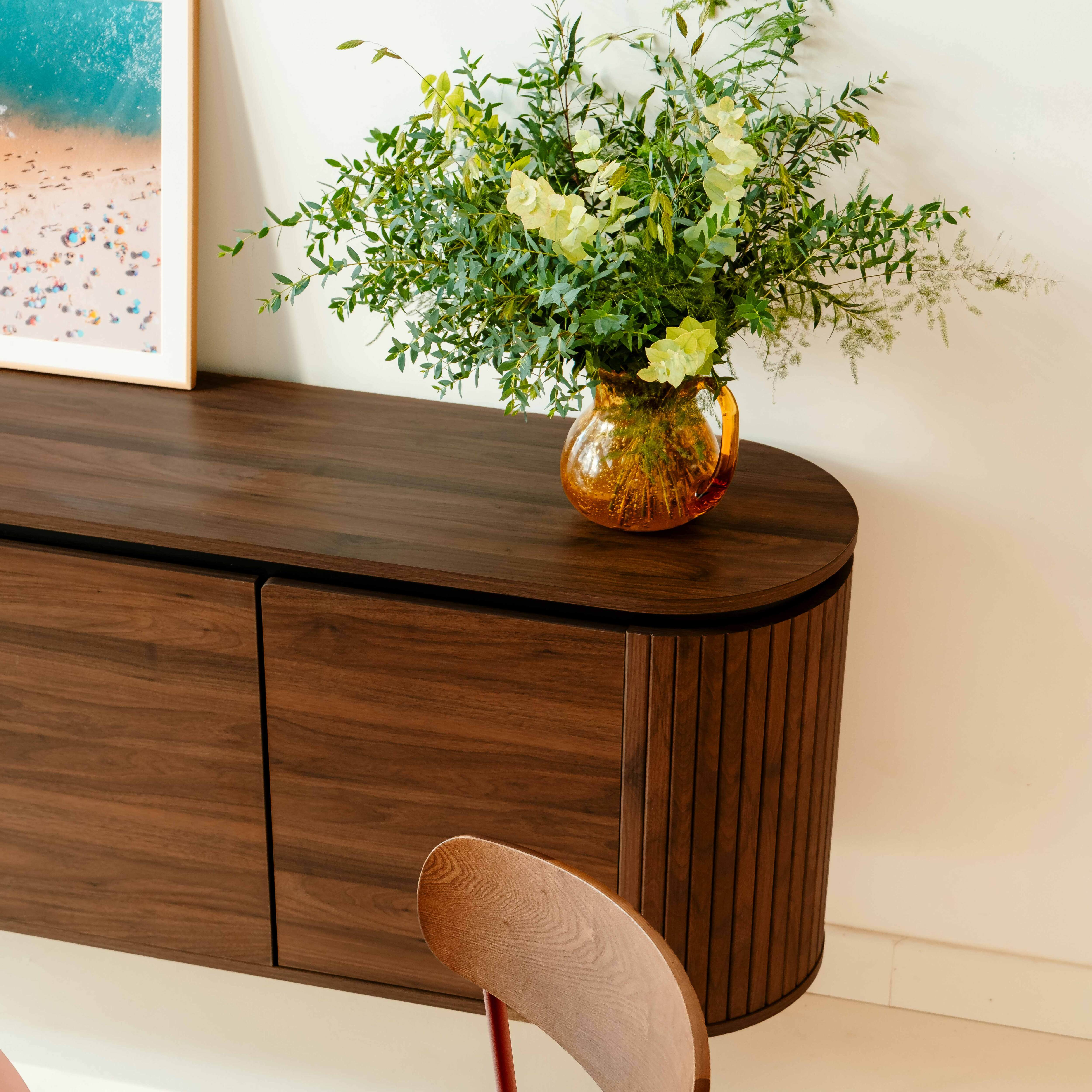 Ein dunkler Holzschrank mit einer Blumenvase darauf. Ein Strandbild hängt an der Wand.