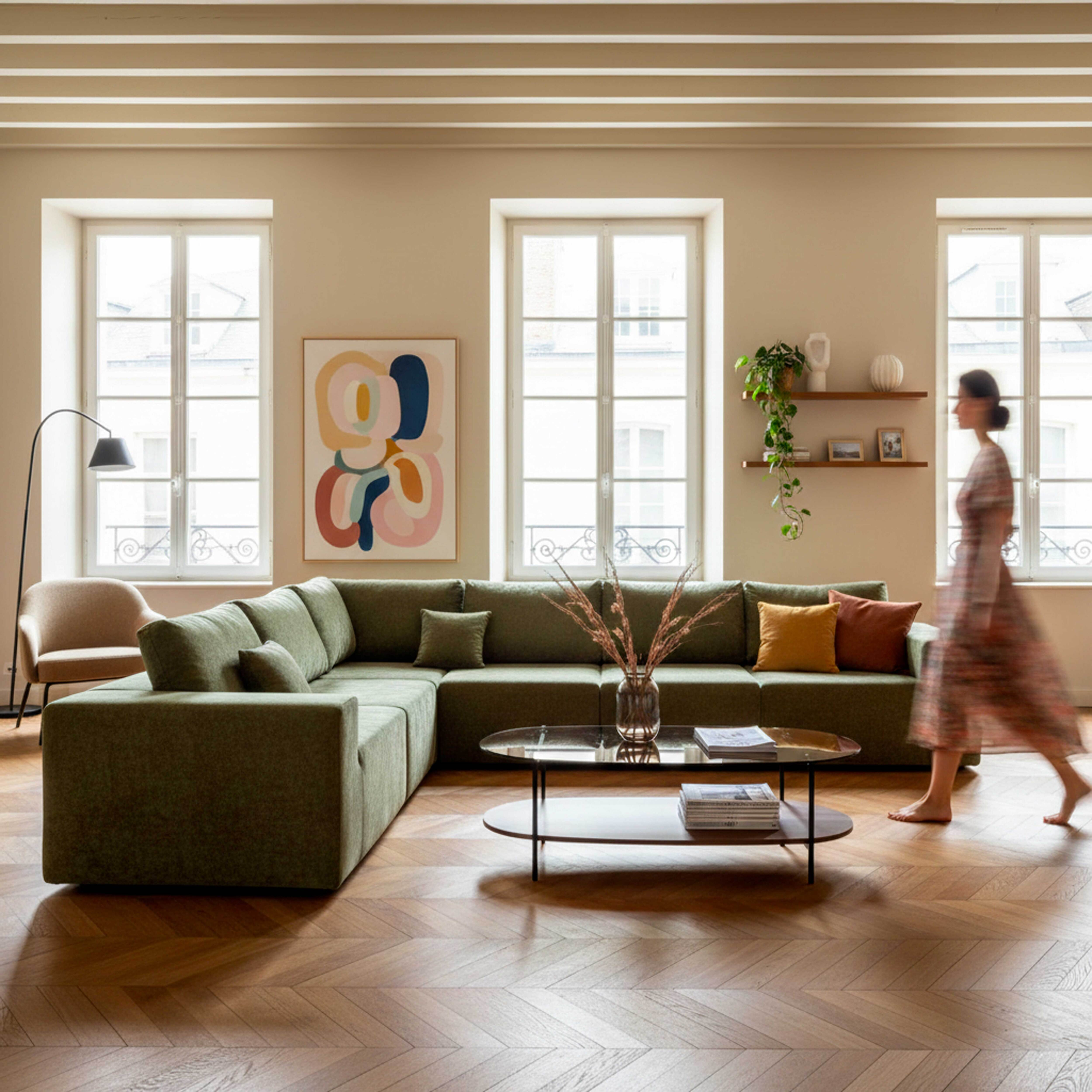 Wohnzimmer mit grünem Sofa, Glastisch, Frau geht, Kunst an der Wand und Fenstern mit Außenansicht.