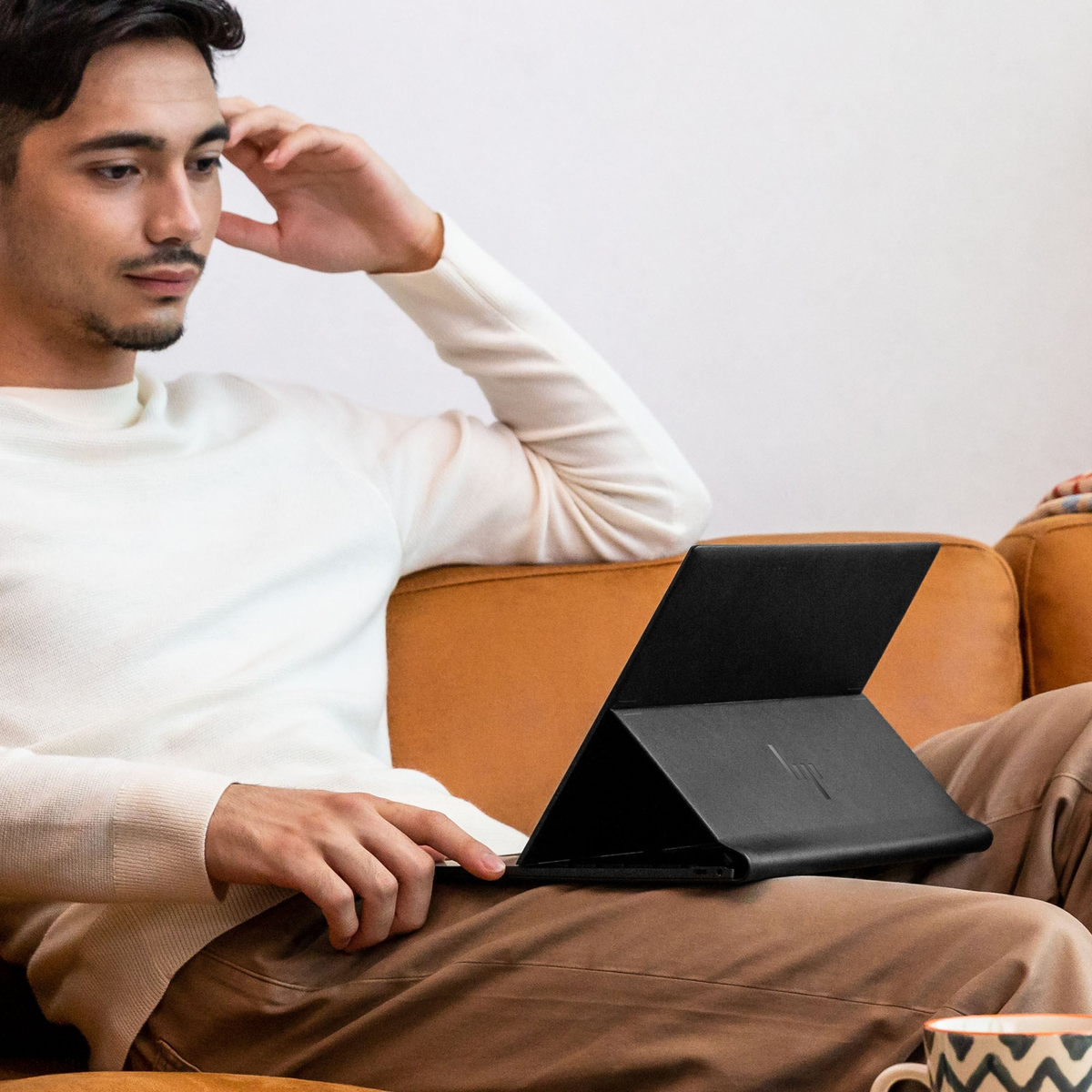 Ein Mann sitzt auf einem Sofa und hält einen schwarzen Laptop. Er trägt einen weißen Pullover und braune Hosen. Der Hintergrund ist weiß.