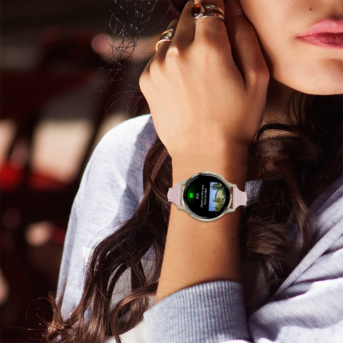 Das Handgelenk einer Frau mit einer Smartwatch. Die Uhr zeigt eine Nachricht an. Sie trägt einen grauen Pullover und hat braune Haare. Sie berührt ihr Gesicht.