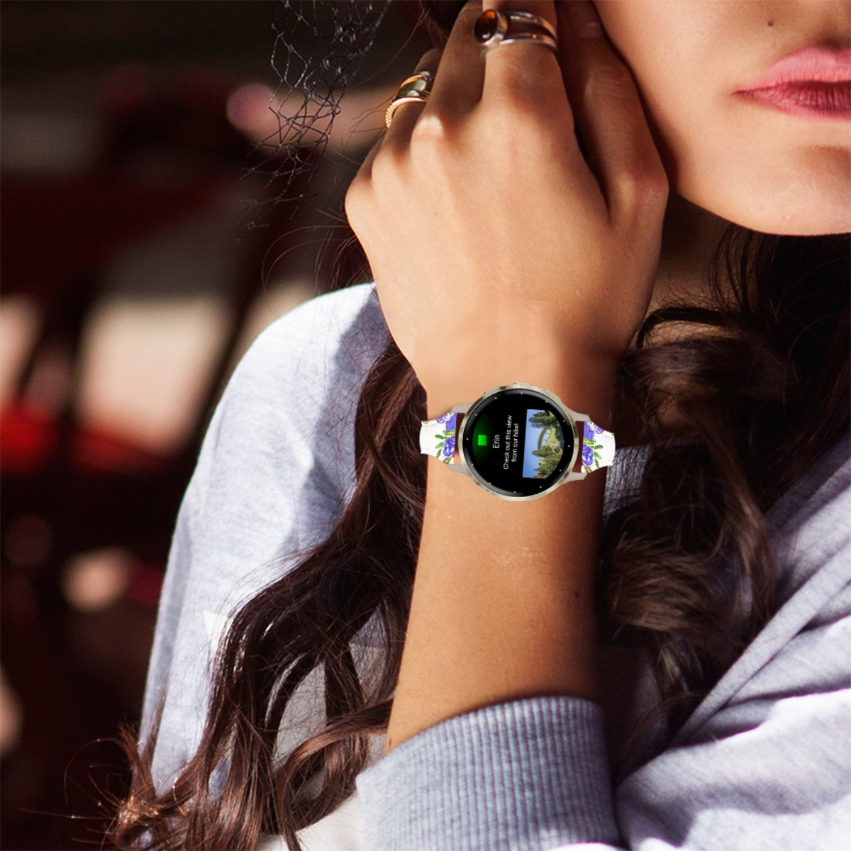 Das Handgelenk einer Frau mit floraler Smartwatch, ihre Hand berührt ihr Gesicht. Sie trägt Ringe. Die Uhr zeigt eine Benachrichtigung.