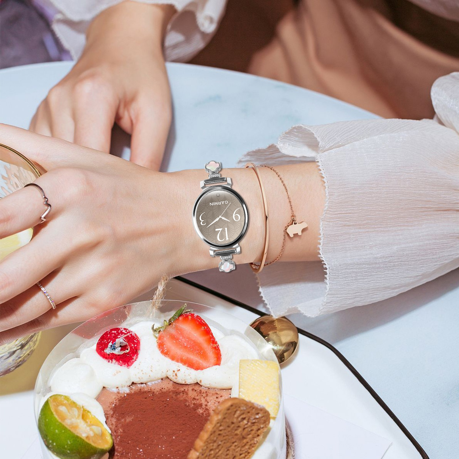 Das Handgelenk einer Frau mit Uhr und Armbändern über einem Kuchen mit Obst. Die Szene ist weich und elegant.