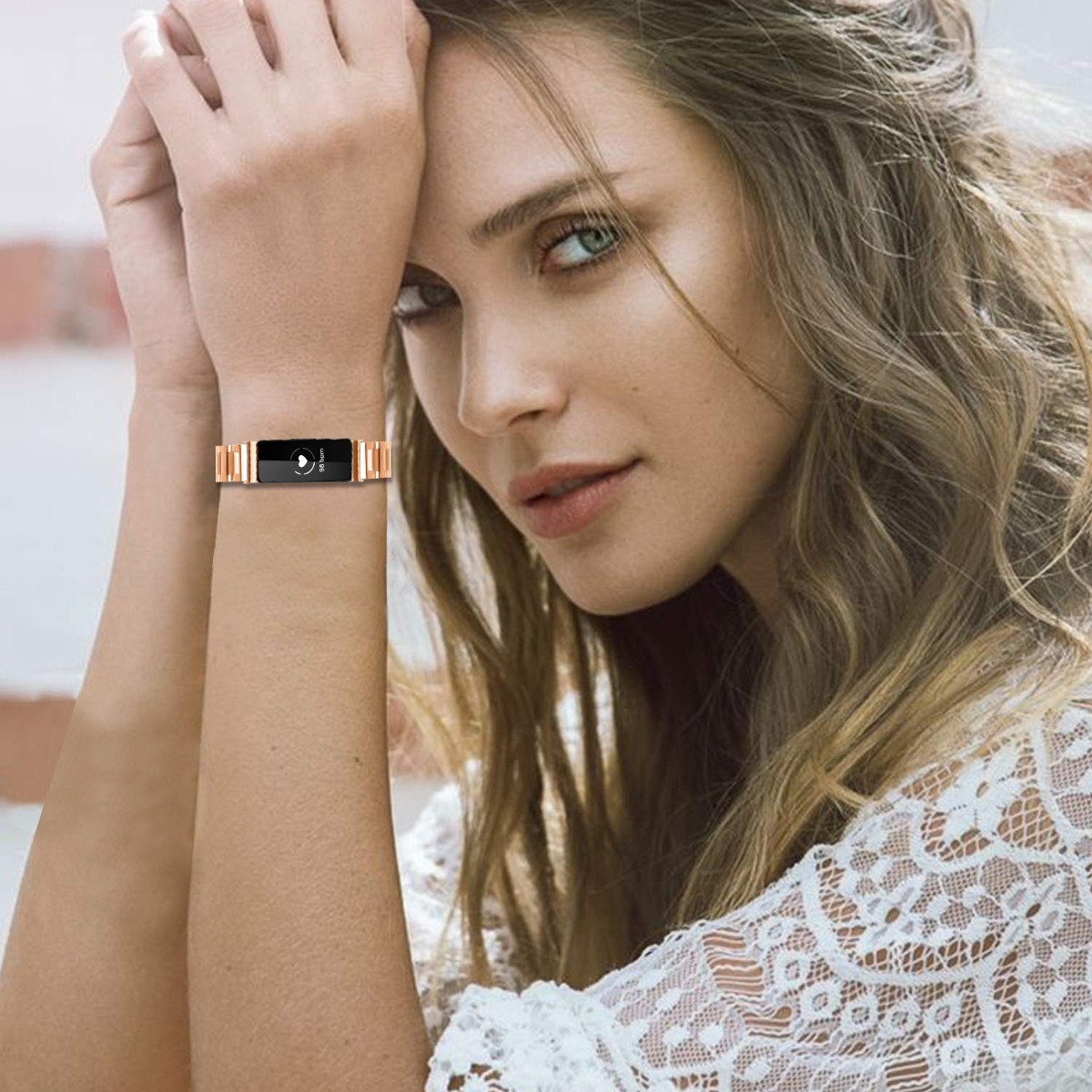 Eine Frau mit langen, welligen Haaren trägt einen Fitnesstracker am Handgelenk. Sie trägt ein weißes Spitzentop und schaut in die Kamera.