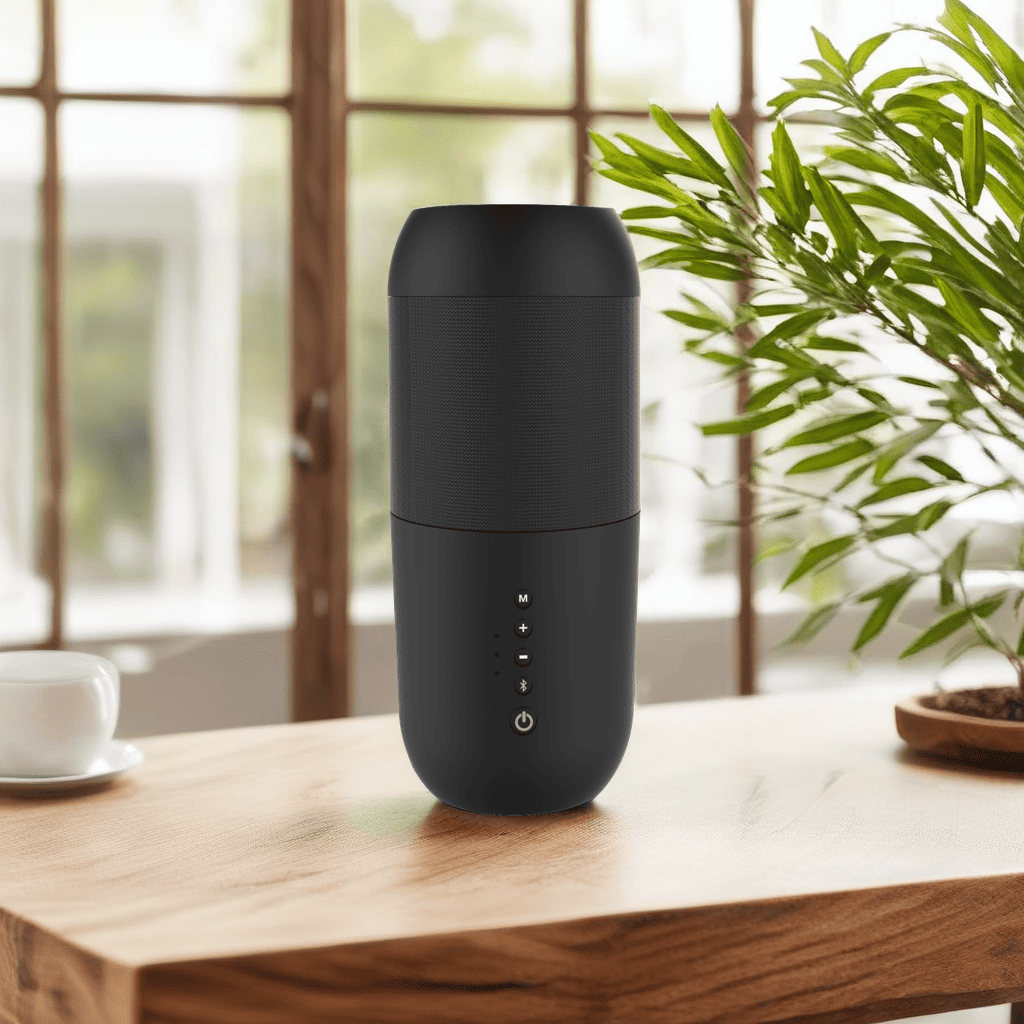 Un altavoz negro sobre una mesa de madera. Una taza, planta y ventana en el fondo. La escena está bien iluminada.