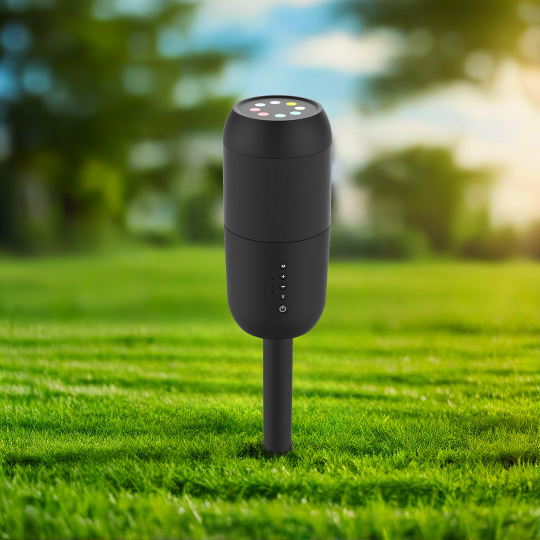 Un altavoz cilíndrico negro con luces de colores en la parte superior, colocado sobre césped verde. El fondo muestra árboles y cielo borrosos.