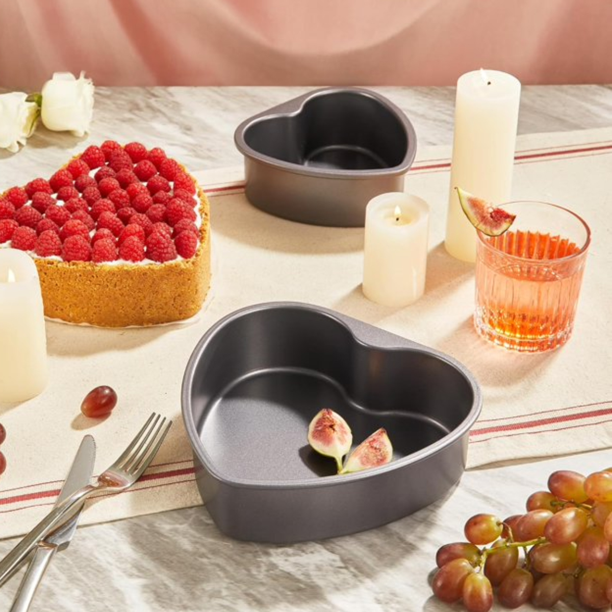 Herzförmiger Kuchen mit Himbeeren, herzförmige Pfannen, Kerzen, Getränk, Feigen, Trauben und Besteck auf einem Tisch.
