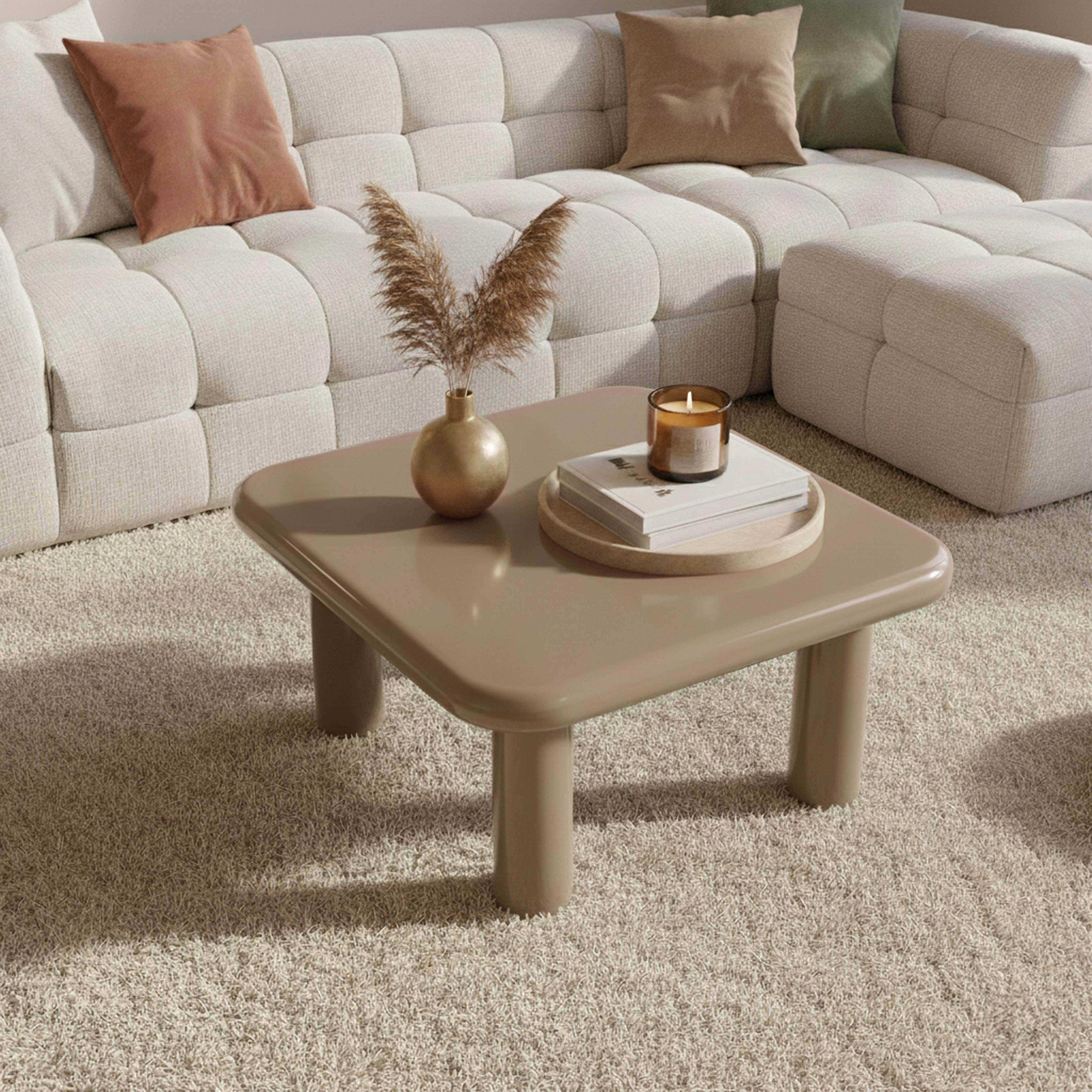 Ein beigefarbener Couchtisch mit Vase und Kerze steht auf einem hellen Teppich, neben einem weißen Sofa mit Kissen.