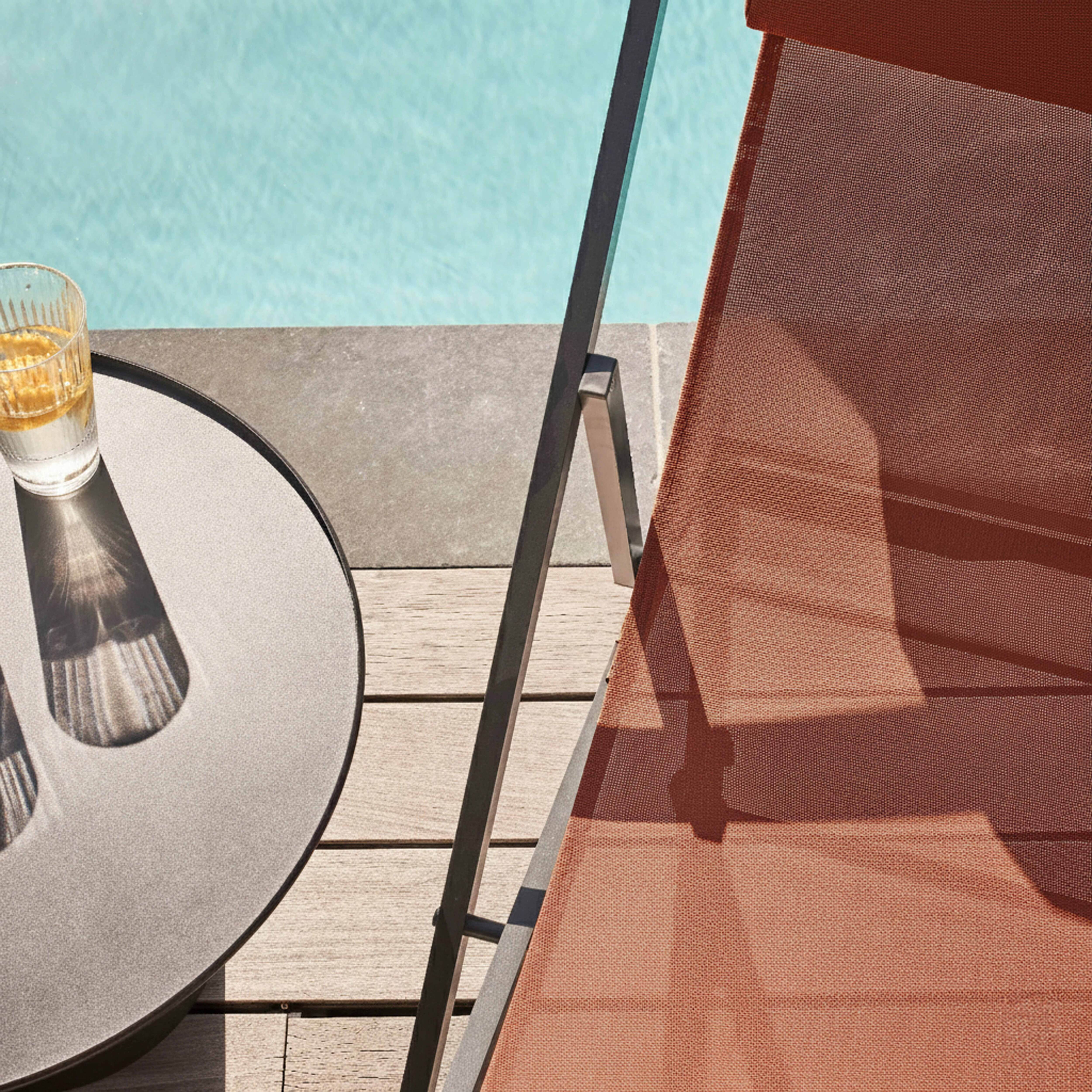 Ein Glas Wasser mit Zitrone auf einem runden Tisch neben einem Pool und einem braunen Stuhl.