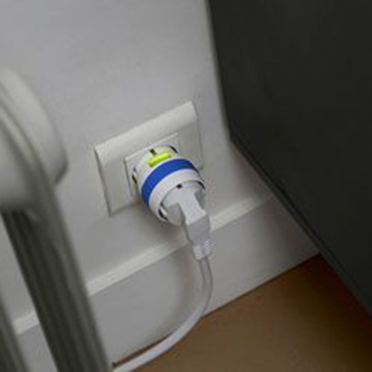 Un enchufe blanco con un adaptador azul y blanco conectado. El cable baja al suelo. Parte de un radiador es visible.