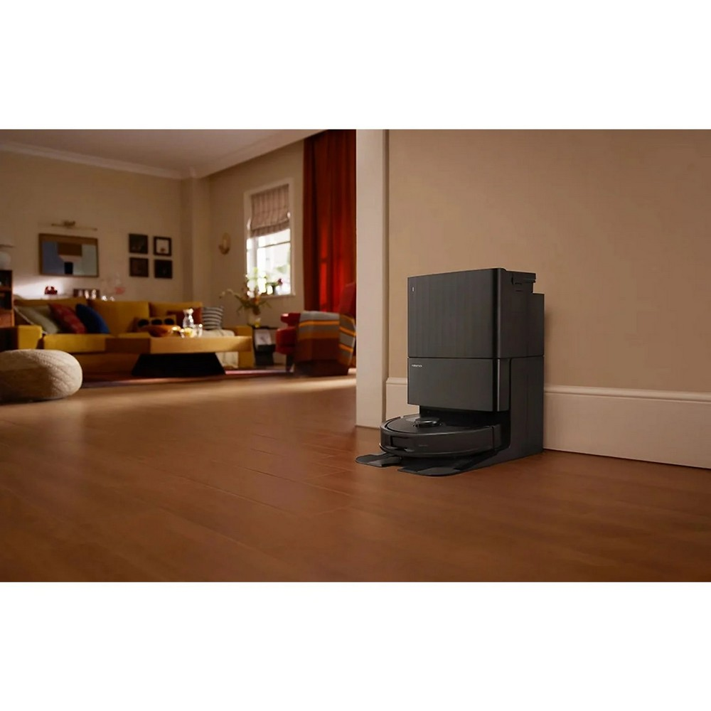 Ein Roboter-Staubsauger und seine Ladestation befinden sich in einem Raum. Der Raum hat Holzböden und eine Couch im Hintergrund.