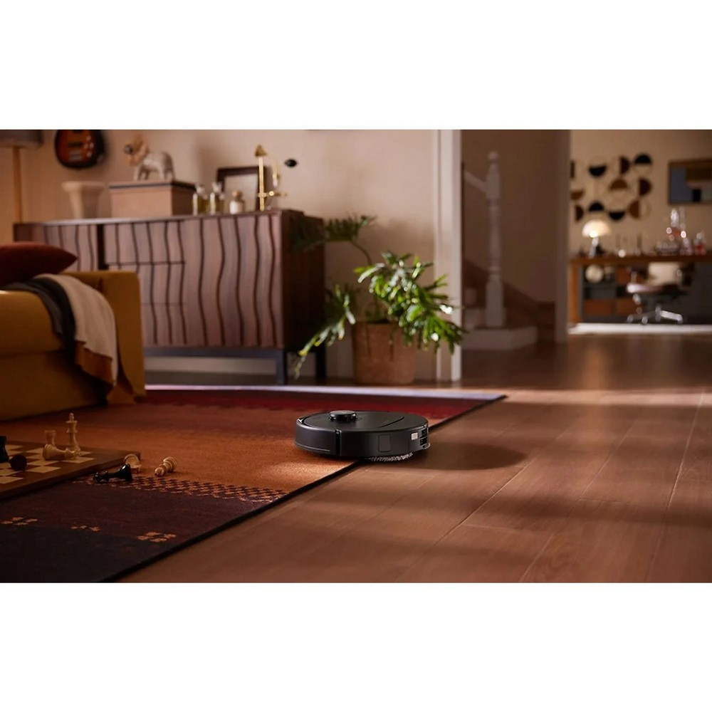 Ein schwarzer Roboter-Staubsauger auf einem Teppich. Links ist ein Schachbrett. Im Hintergrund ein Sofa und ein Schrank.