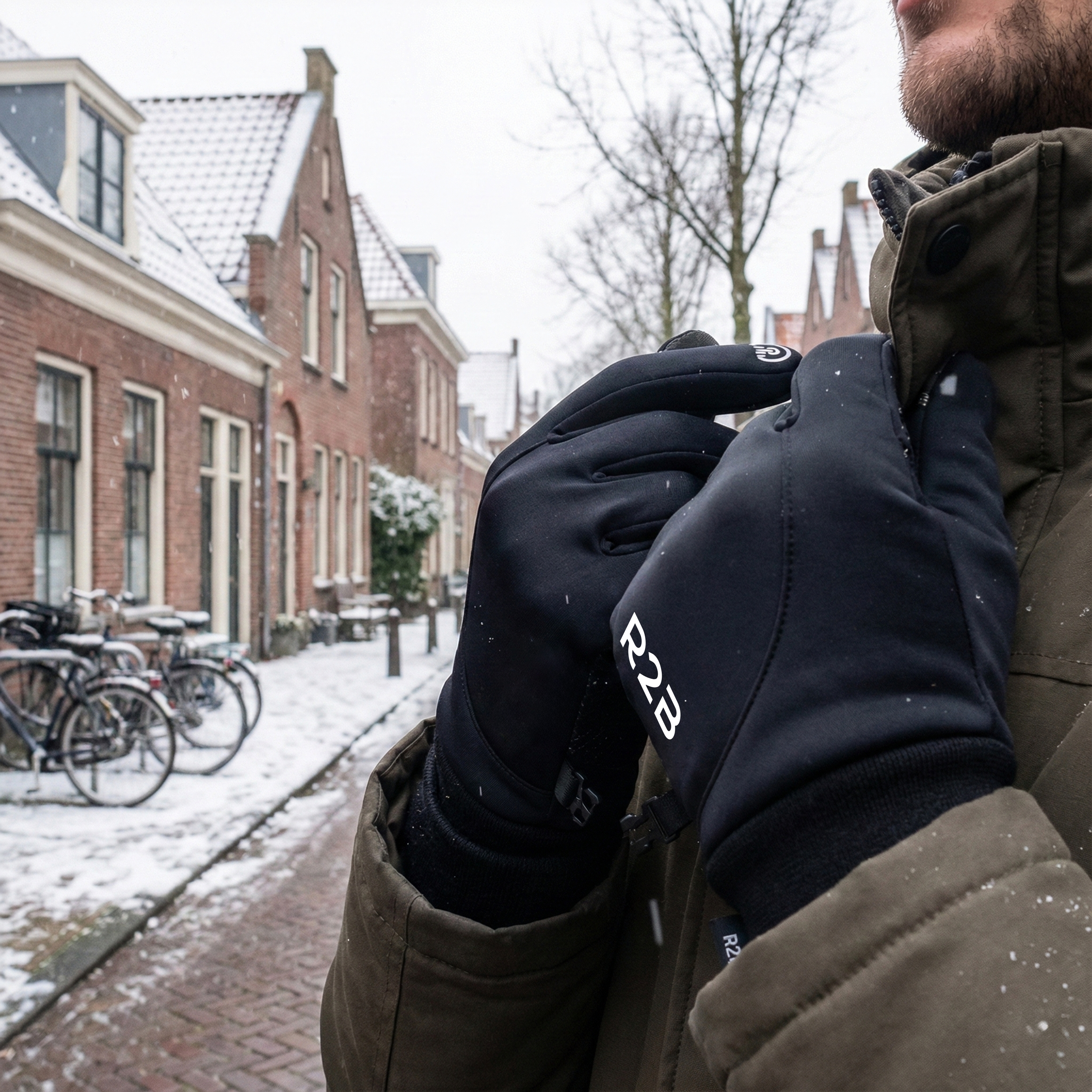 Personne portant des gants noirs avec le logo R2B ajuste le col de son manteau dans une rue enneigée.