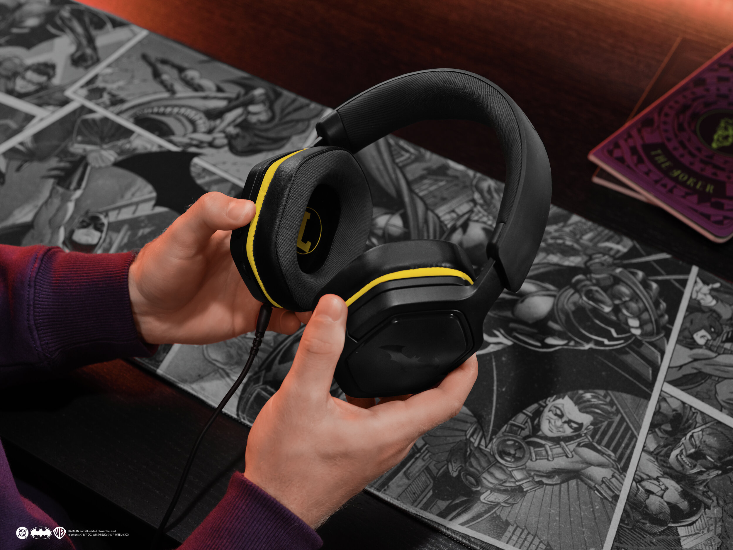 Osoba trzyma czarne słuchawki z żółtymi akcentami. Widoczne jest tło komiksu o Batmanie.