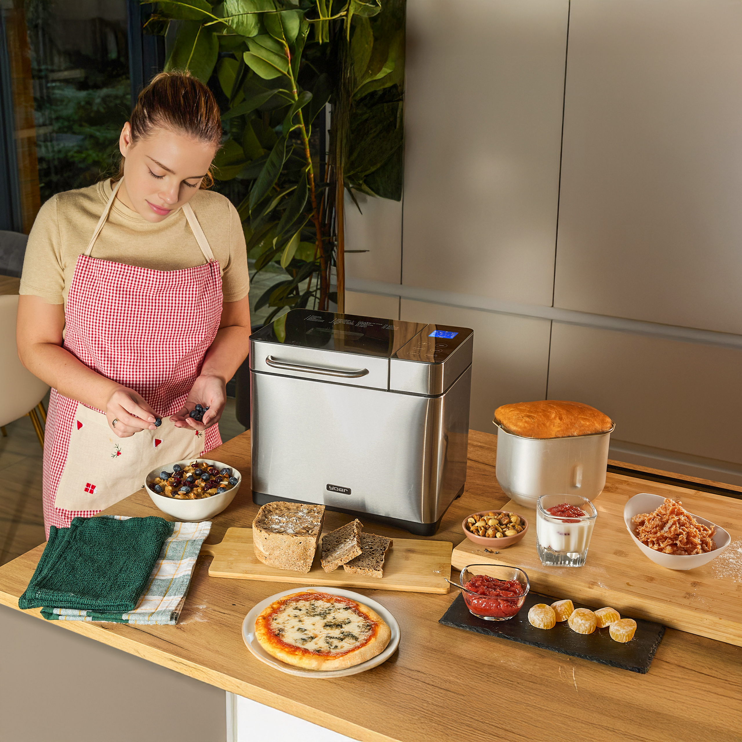 Kobieta w fartuchu przygotowuje jedzenie na blacie z maszyną do chleba, chlebem, pizzą i różnymi składnikami.