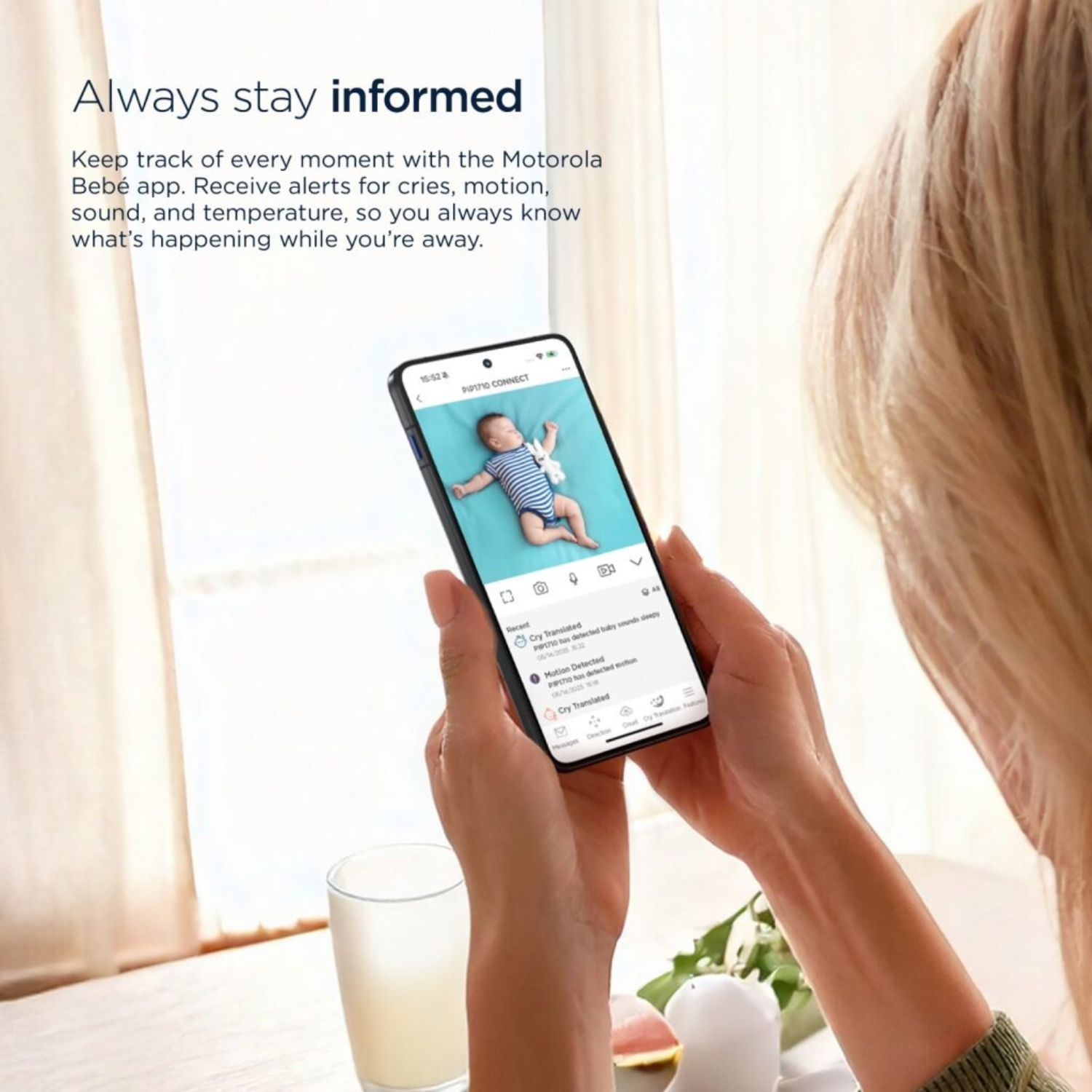 Frau hält Telefon mit Babyfon-App. Der Bildschirm zeigt ein Baby und Benachrichtigungen. Ein Glas und ein Licht sind im Bild.