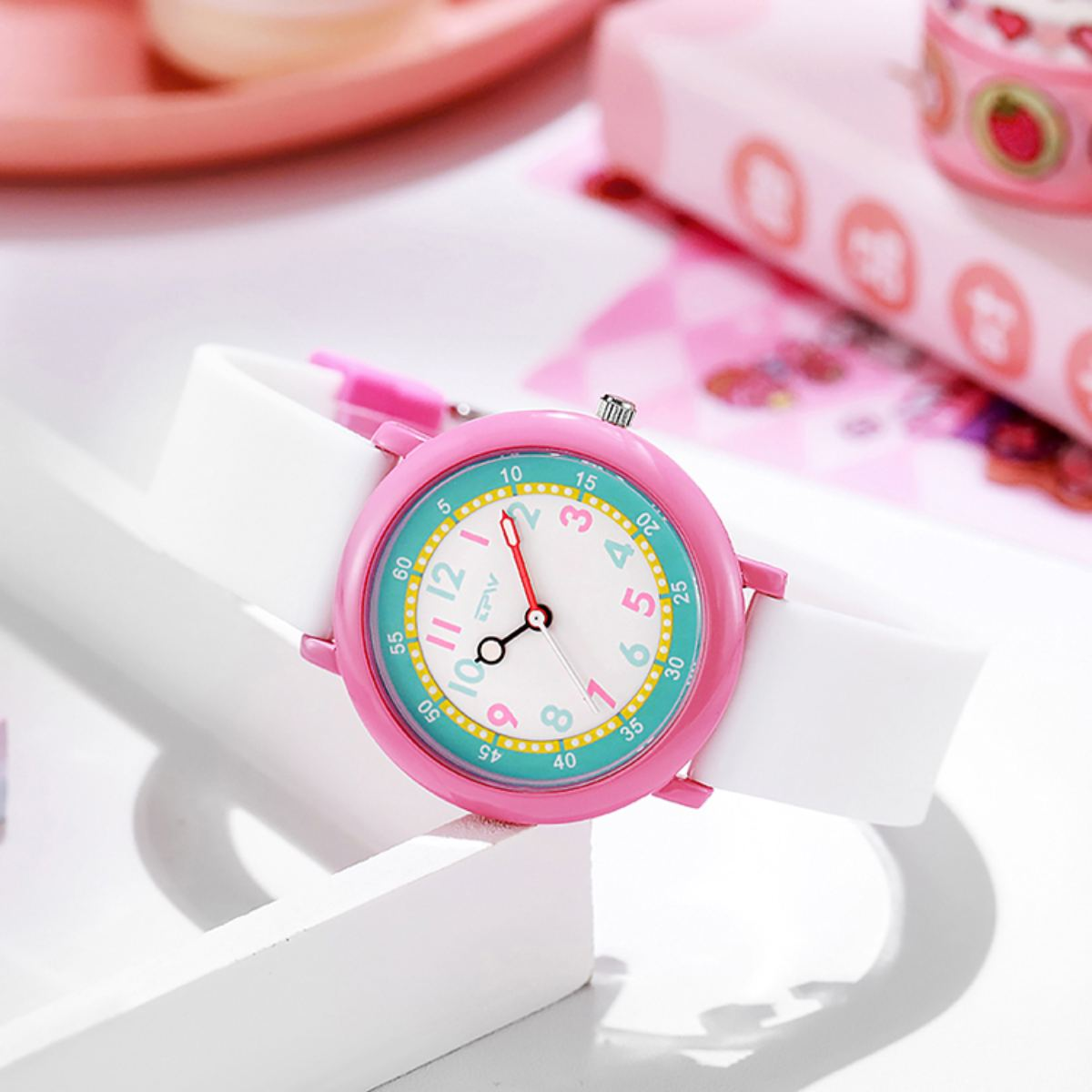 Eine rosa Uhr mit weißem Band liegt auf einer weißen Oberfläche. Das Zifferblatt zeigt Zahlen und Zeiger an.