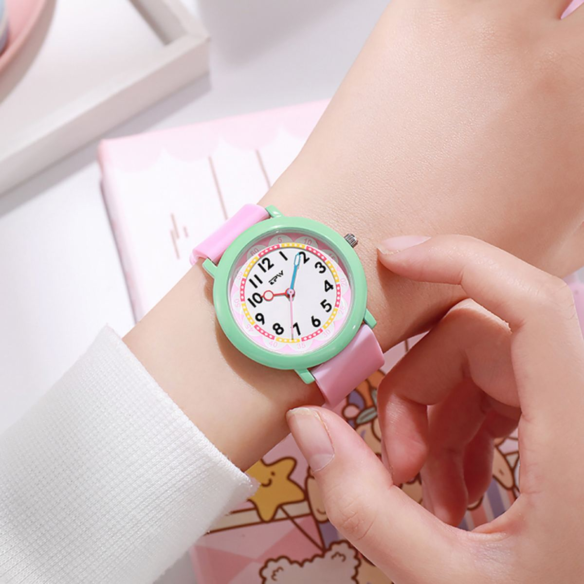 Una persona indossa un orologio rosa e verde. L'orologio ha un quadrante bianco con numeri neri. Lo sfondo include un quaderno rosa.