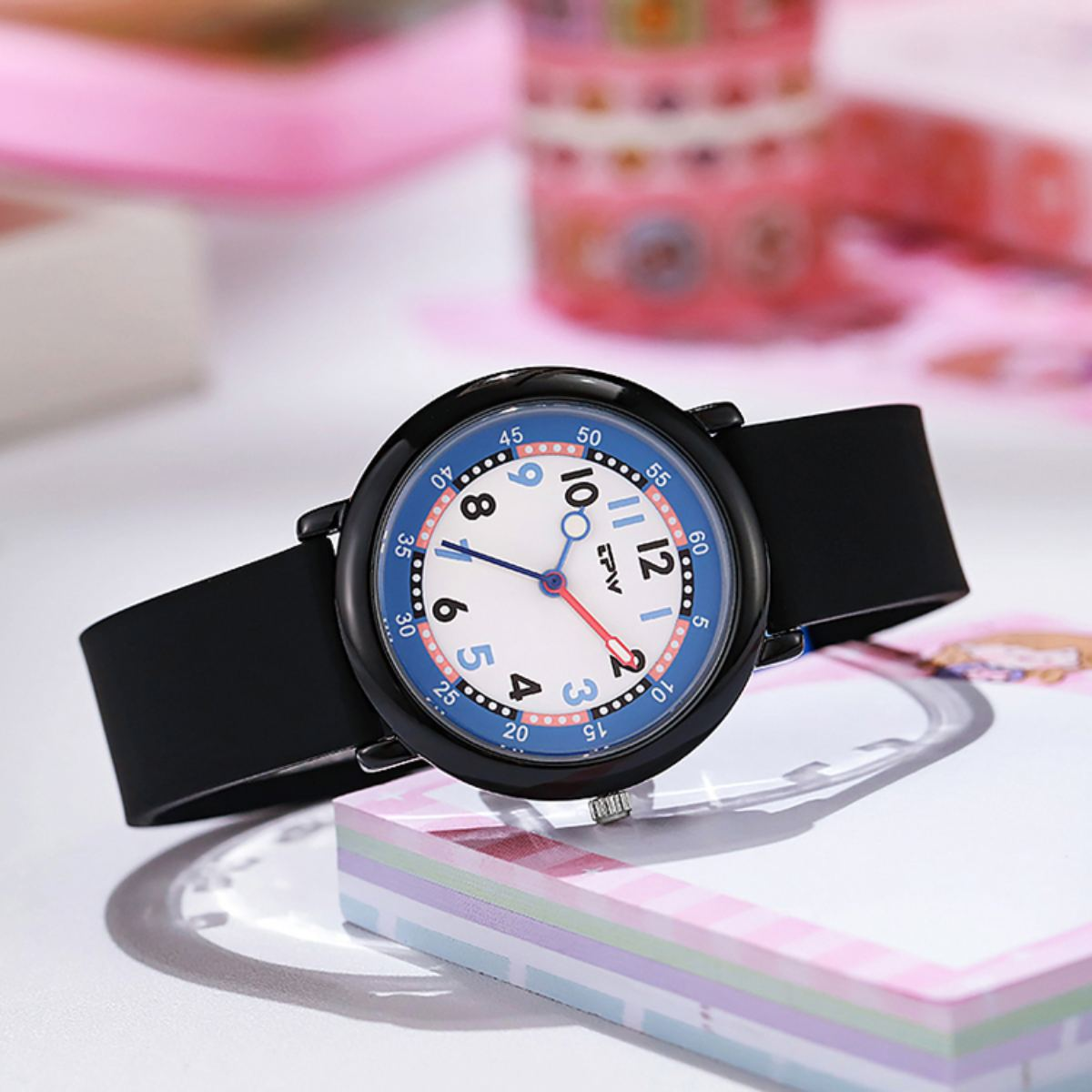 Eine schwarze Armbanduhr mit weißem und blauem Zifferblatt. Die Uhr zeigt die Uhrzeit an. Sie liegt auf einer weißen Oberfläche mit anderen Gegenständen im Hintergrund.