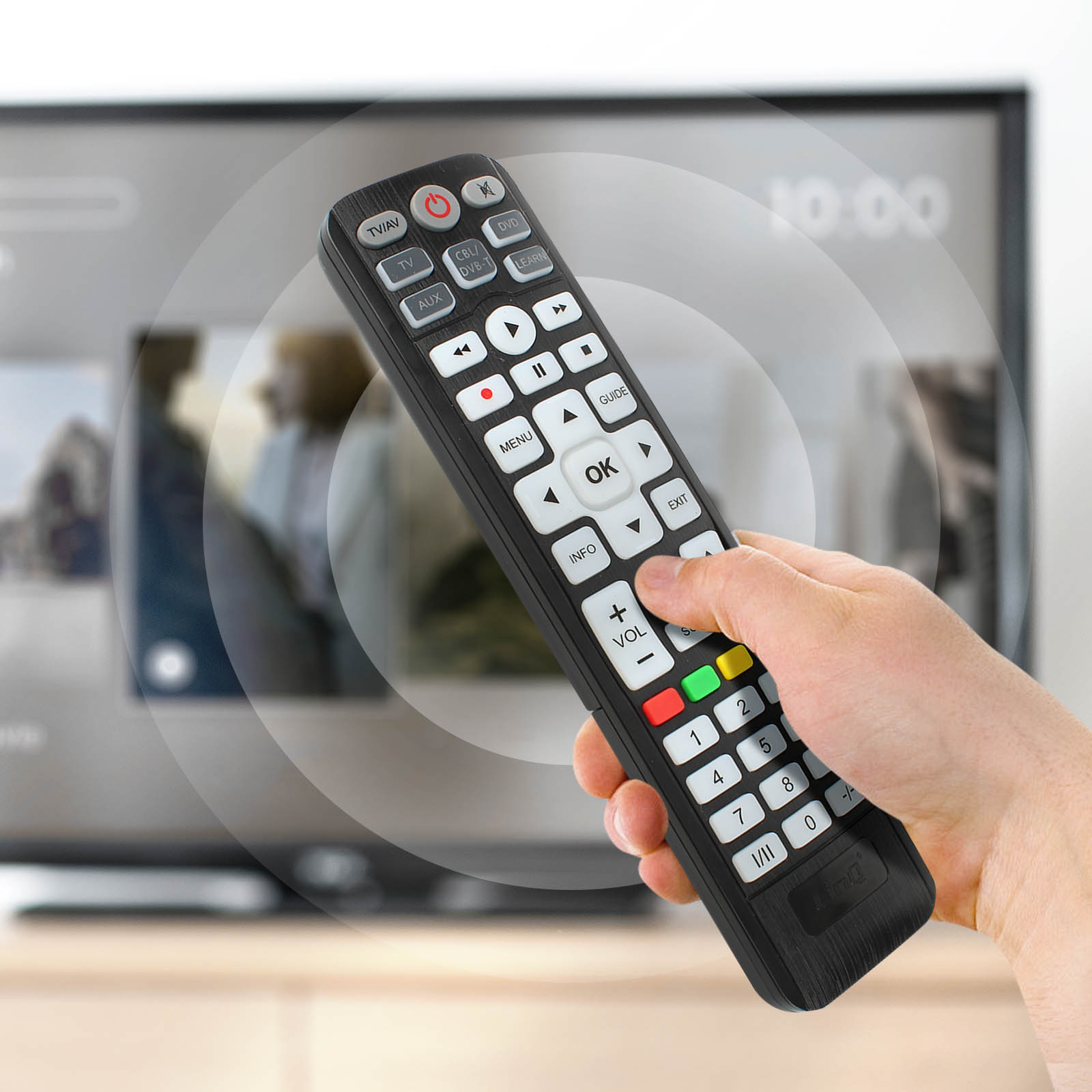 Una mano sostiene un mando a distancia negro, pulsando un botón. Una pantalla de TV está en el fondo.