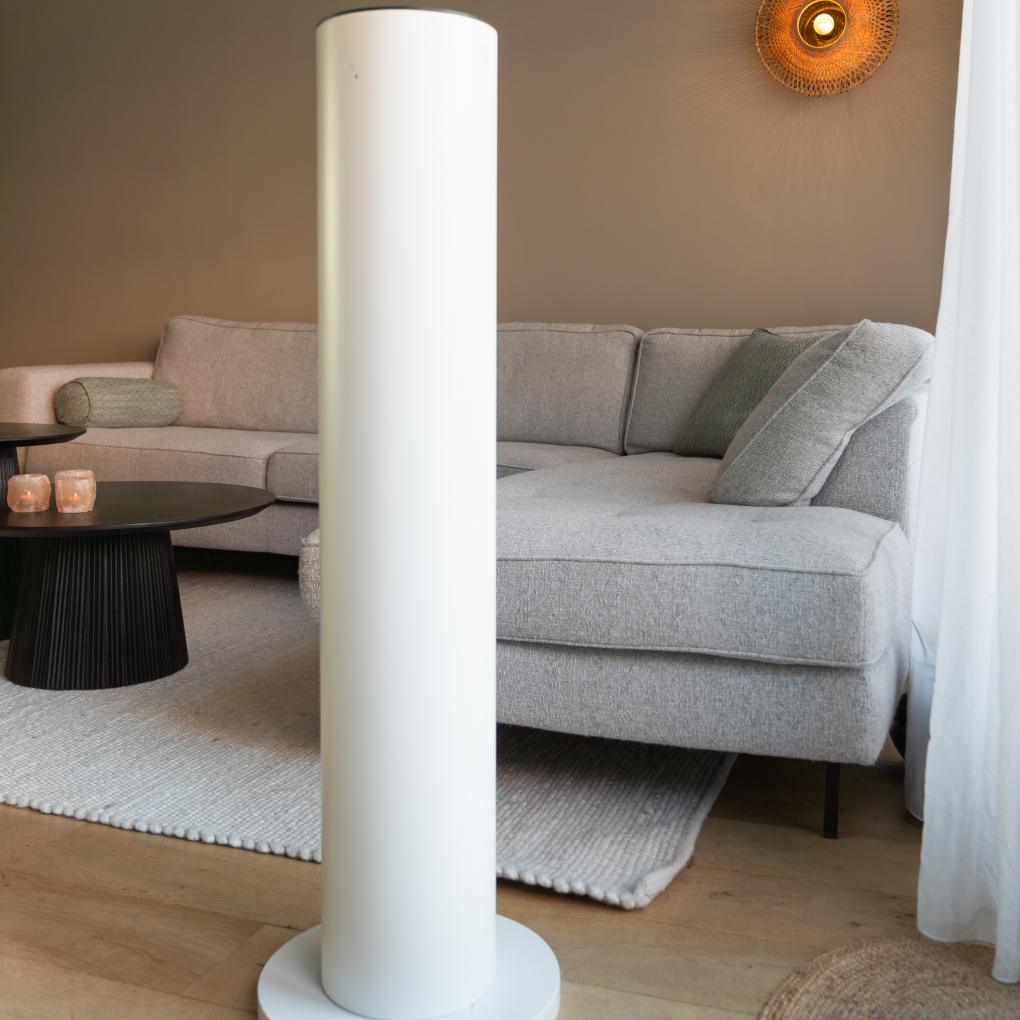 Wit cilindrisch object in een woonkamer met een grijze bank, zwarte tafel en een decoratieve wandlamp. Lichte houten vloer.