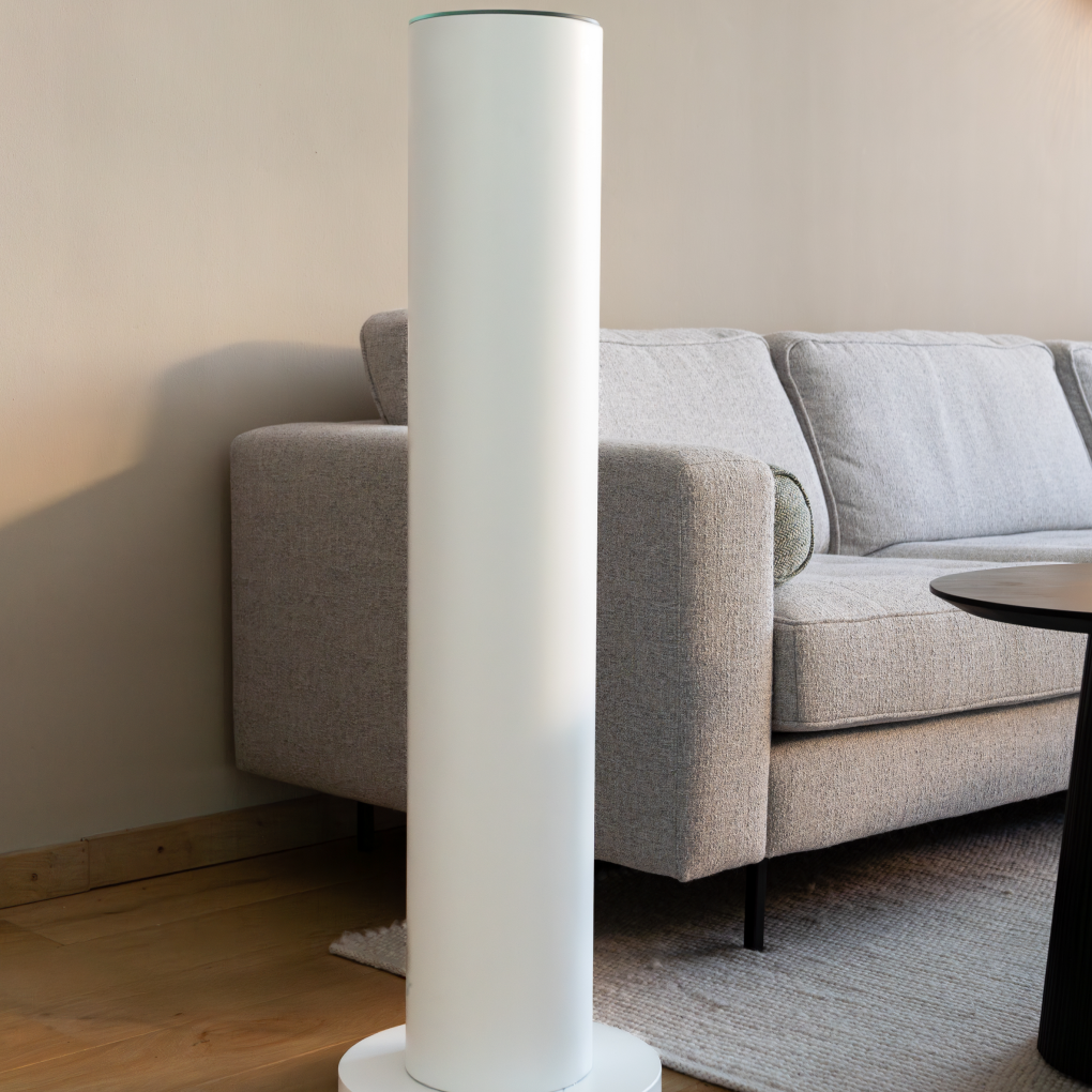 Een witte cilindrische luchtreiniger staat voor een grijze bank en een beige muur. Een tapijt en een zwarte tafel zijn ook zichtbaar.