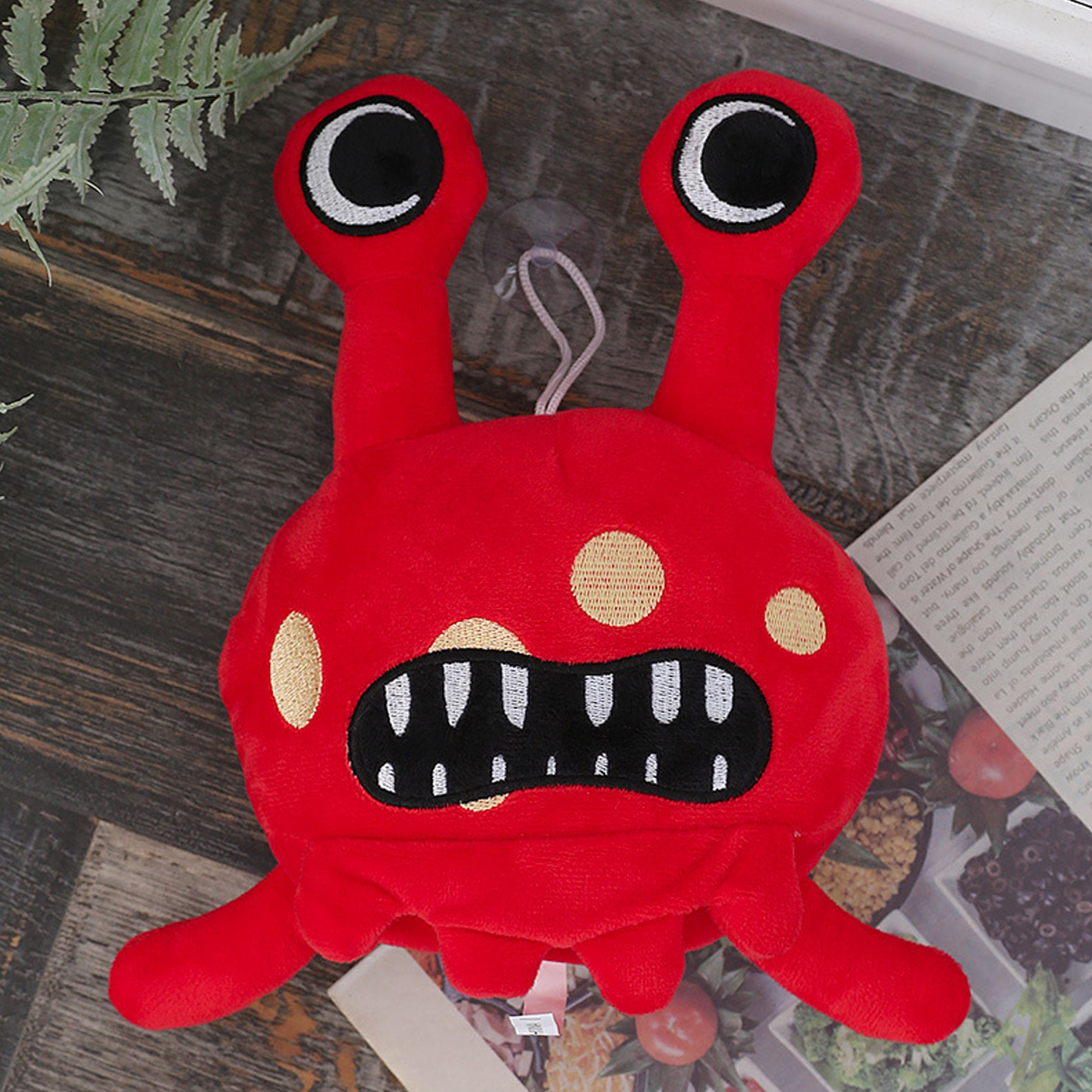 Ein rotes Plüschmonster-Spielzeug mit großen Augen, Zähnen und Punkten, platziert auf einer Holzoberfläche mit Farn und Papier.
