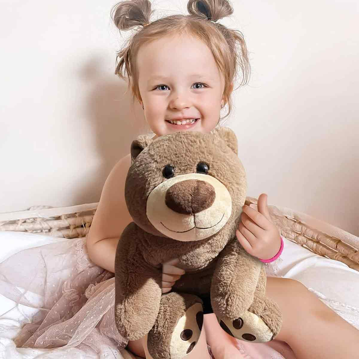 Ein lächelndes Kind hält einen braunen Teddybären. Das Kind hat blonde Zöpfe und trägt ein rosa Armband. Sie sitzen auf einem Bett.