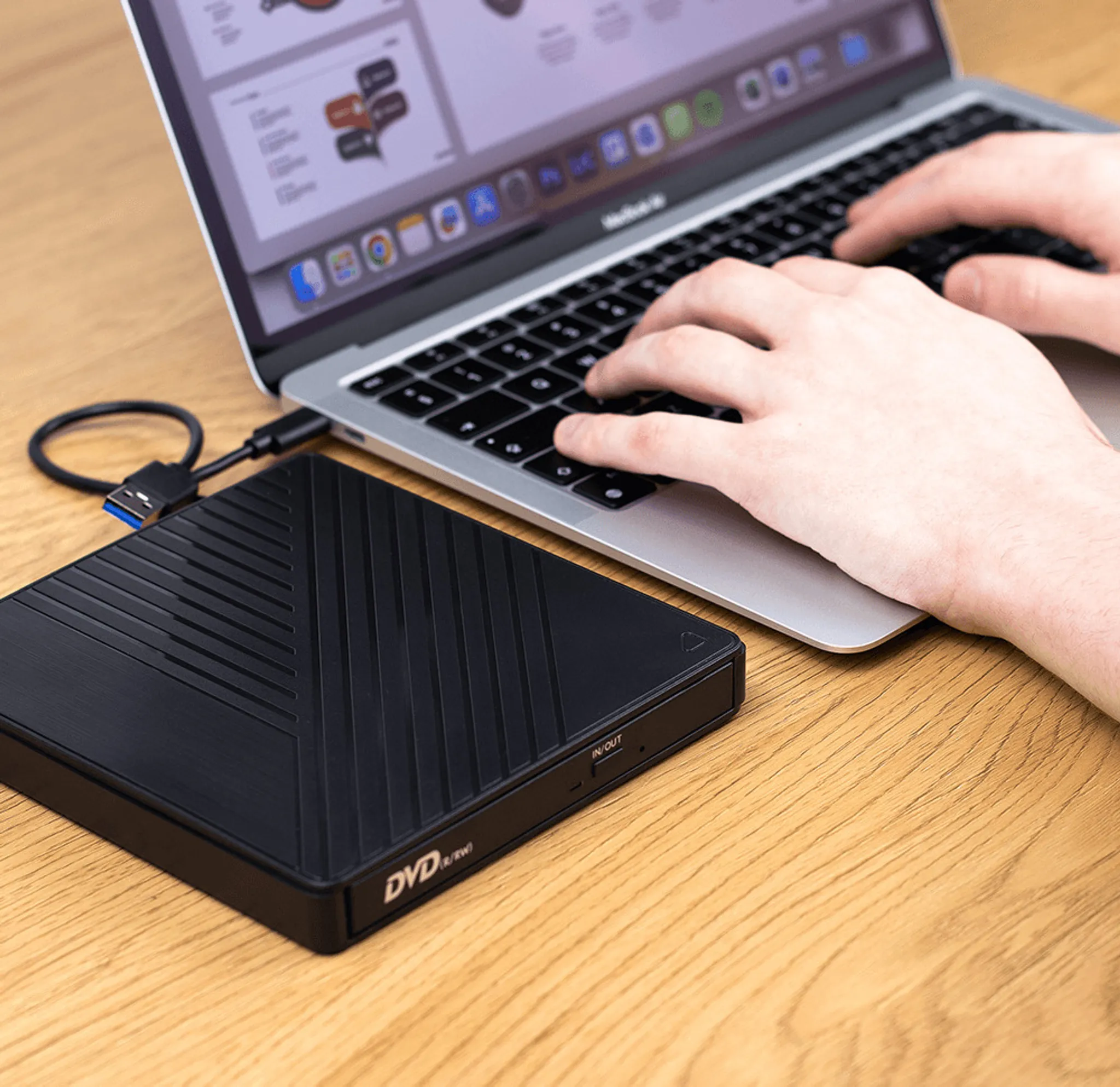 Ein Laptop ist mit einem schwarzen externen DVD-Laufwerk verbunden. Hände tippen auf der Laptop-Tastatur auf einem Holztisch.