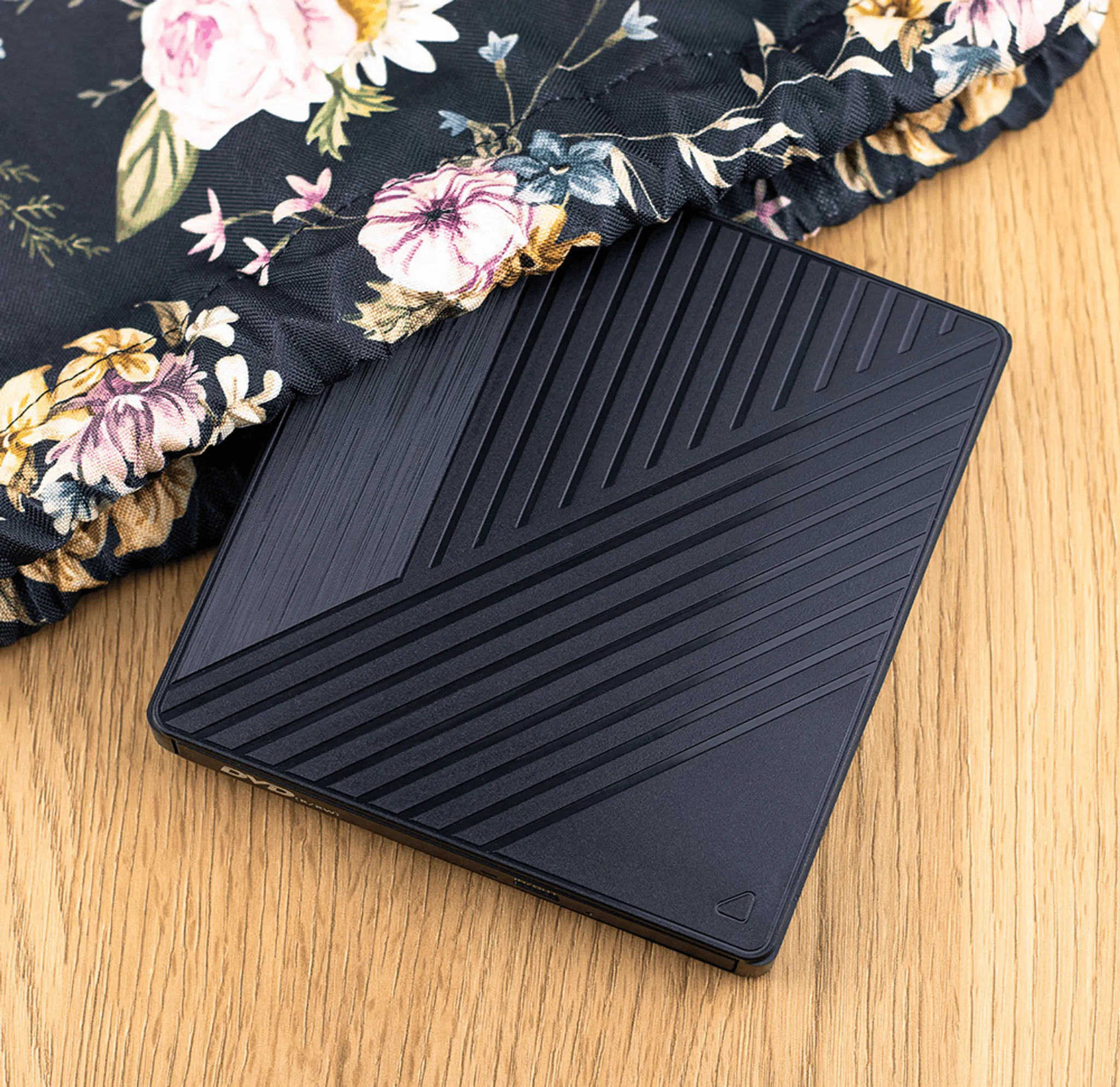 Ein schwarzes Gerät mit diagonalen Linien liegt auf Holz. Ein Blumengewebe mit rosa Blüten liegt oben.