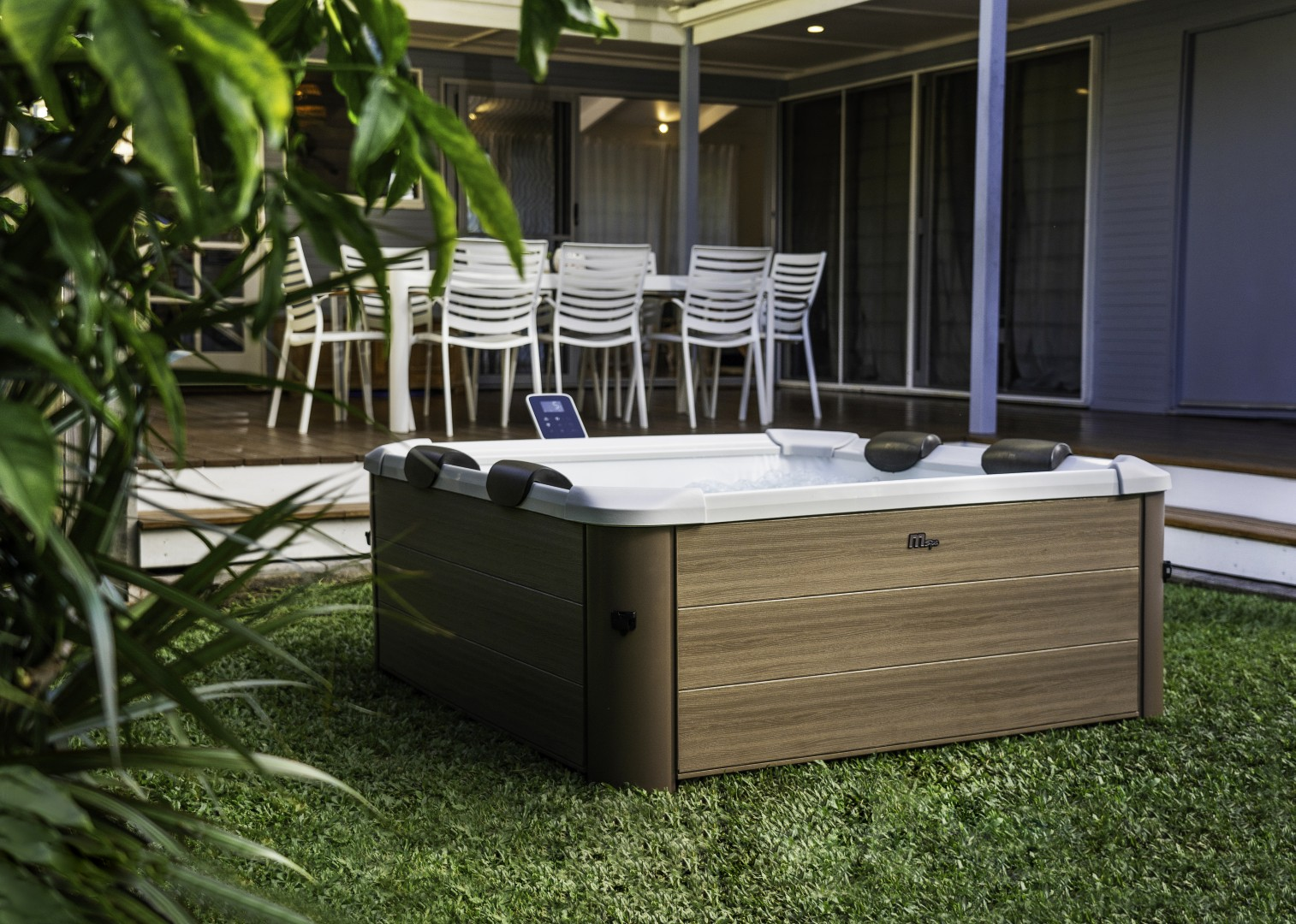 Ein Whirlpool auf Rasen, hinter grünen Pflanzen. Im Hintergrund stehen weiße Stühle und ein Tisch auf einem Deck.