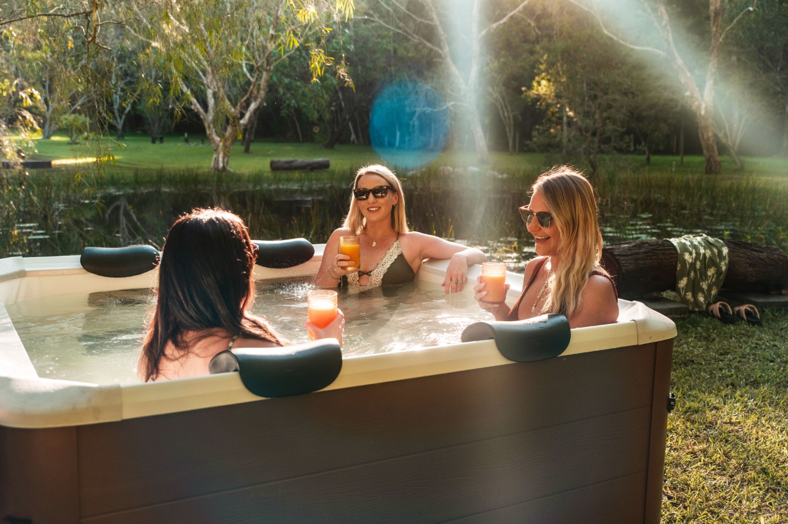 Drei Frauen in einem Whirlpool, die Getränke halten. Sonniger Außenbereich mit Bäumen und einem kleinen Teich im Hintergrund.