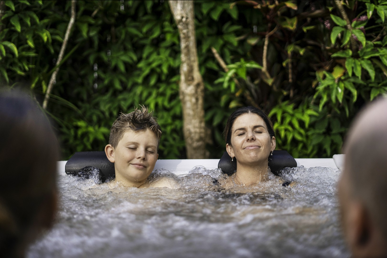 Eine Frau und ein Junge entspannen sich im Whirlpool, Augen geschlossen. Grünes Laub im Hintergrund, zwei verschwommene Personen an den Seiten.