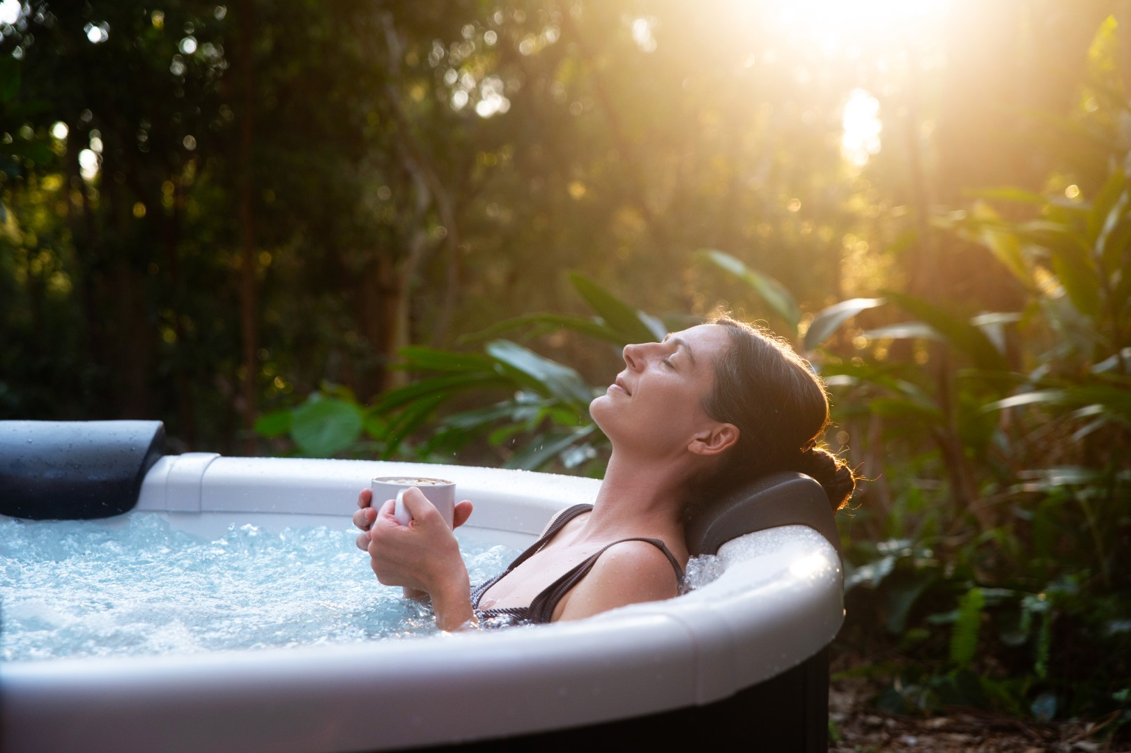 Eine Frau entspannt sich in einem Whirlpool und hält eine Tasse. Ihre Augen sind geschlossen, mit Sonnenlicht im Hintergrund. Grünes Laub.